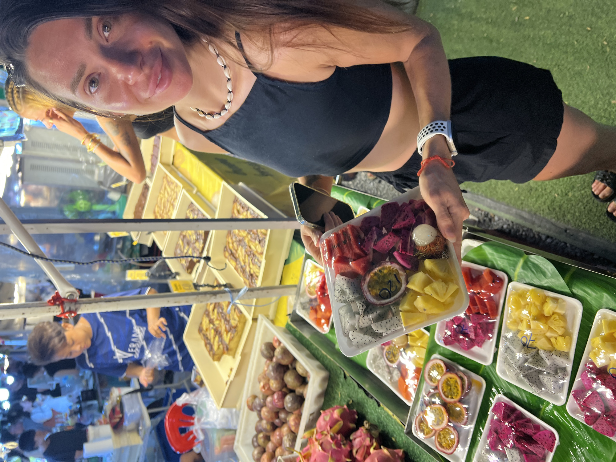A woman stands at a fruit stand in Baan Bo Phut, Thailand. She holds a tray of fresh fruit including dragon fruit, watermelon, pineapple, passionfruit, and lychee, while wearing a black top and shorts. The stand offers a variety of fruits like dragon fruit and passion fruit, and trays of pastries. A vendor in a blue shirt stands behind the stall, where several people are gathered. The scene is brightly lit, suggesting an evening market setting.