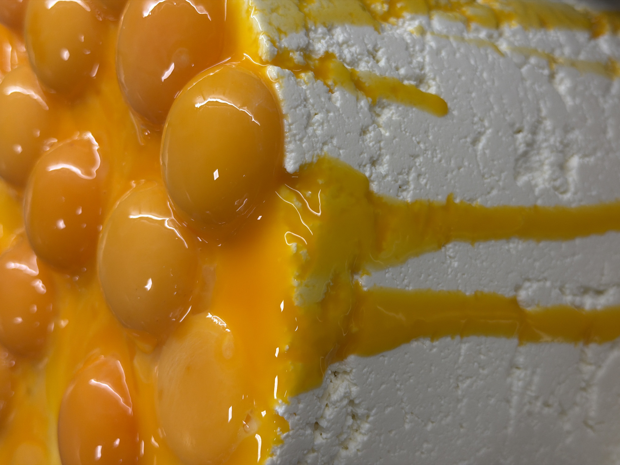 The image shows a close-up of food. On the left side, there are many unbroken egg yolks covered in a thin layer of translucent, viscous fluid, presumably egg white. On the right side, there is a block of white, porous solid material, likely a type of cheese. Yellow fluid runs across the cheese's surface in several streams. The environment is indoor, with artificial lighting creating highlights on the yolks. The image does not depict any people or recognizable city elements, so the location in Belgrade, Serbia, cannot be confirmed or denied.