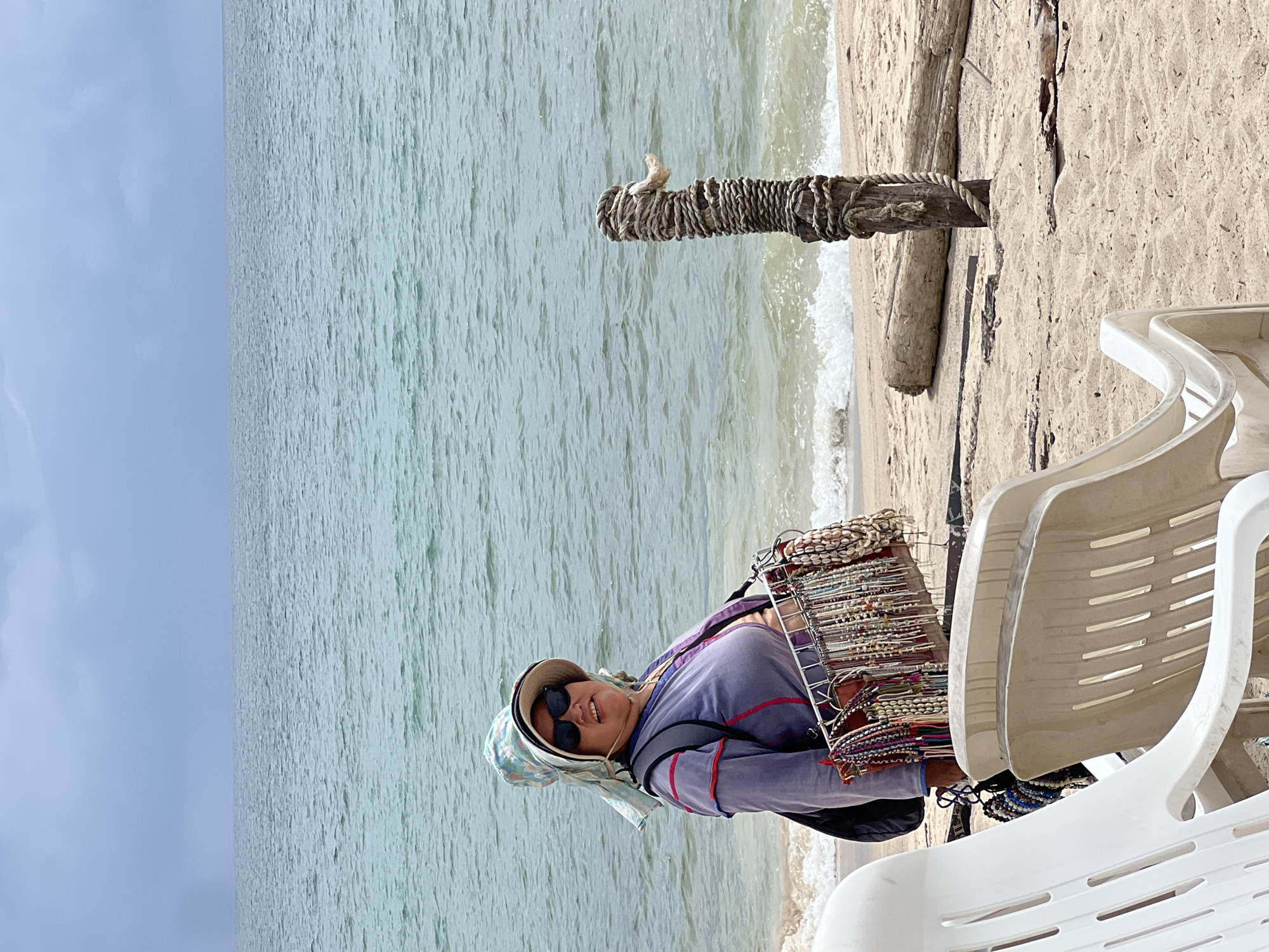A woman stands on a sandy beach, facing the viewer. She is wearing sunglasses, a wide-brimmed hat, and a light purple long-sleeved shirt. She has a display rack of beaded necklaces and bracelets hanging from a harness worn on her chest. The rack is filled with various colorful jewelry, including some made with shells. Behind her, the ocean stretches out to the horizon under a cloudy sky. In the foreground, two empty beach chairs are visible. To the right of the woman, a wooden post is wrapped with thick rope, and a large piece of driftwood lies on the sand behind it. The scene appears to be a commercial setting on a beach, likely in a tourist area.