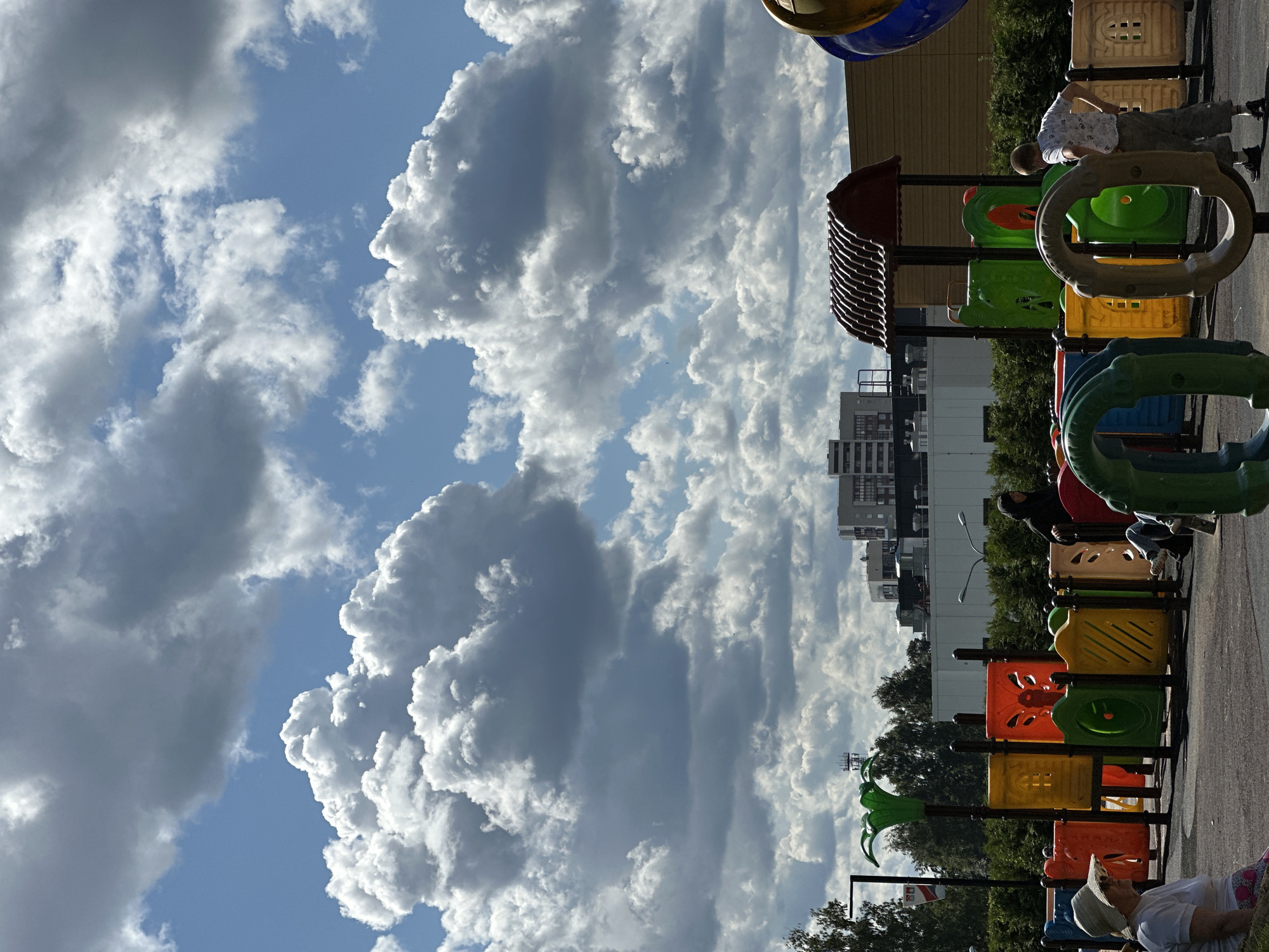 The image depicts a playground in Vsevolozhsk, Russia, under a dramatic sky filled with large, fluffy white and gray clouds. In the foreground, a woman wearing a wide-brimmed hat sits on a dark surface, looking to her left. Adjacent to her is a colorful playground structure with sections in red, yellow, and green, featuring intricate designs. Further back, a boy in a patterned white t-shirt and gray pants stands with his hands on his hips, facing away from the camera. Behind the playground, buildings and trees form the urban landscape. The overall atmosphere is one of a bright but overcast day at a public park.