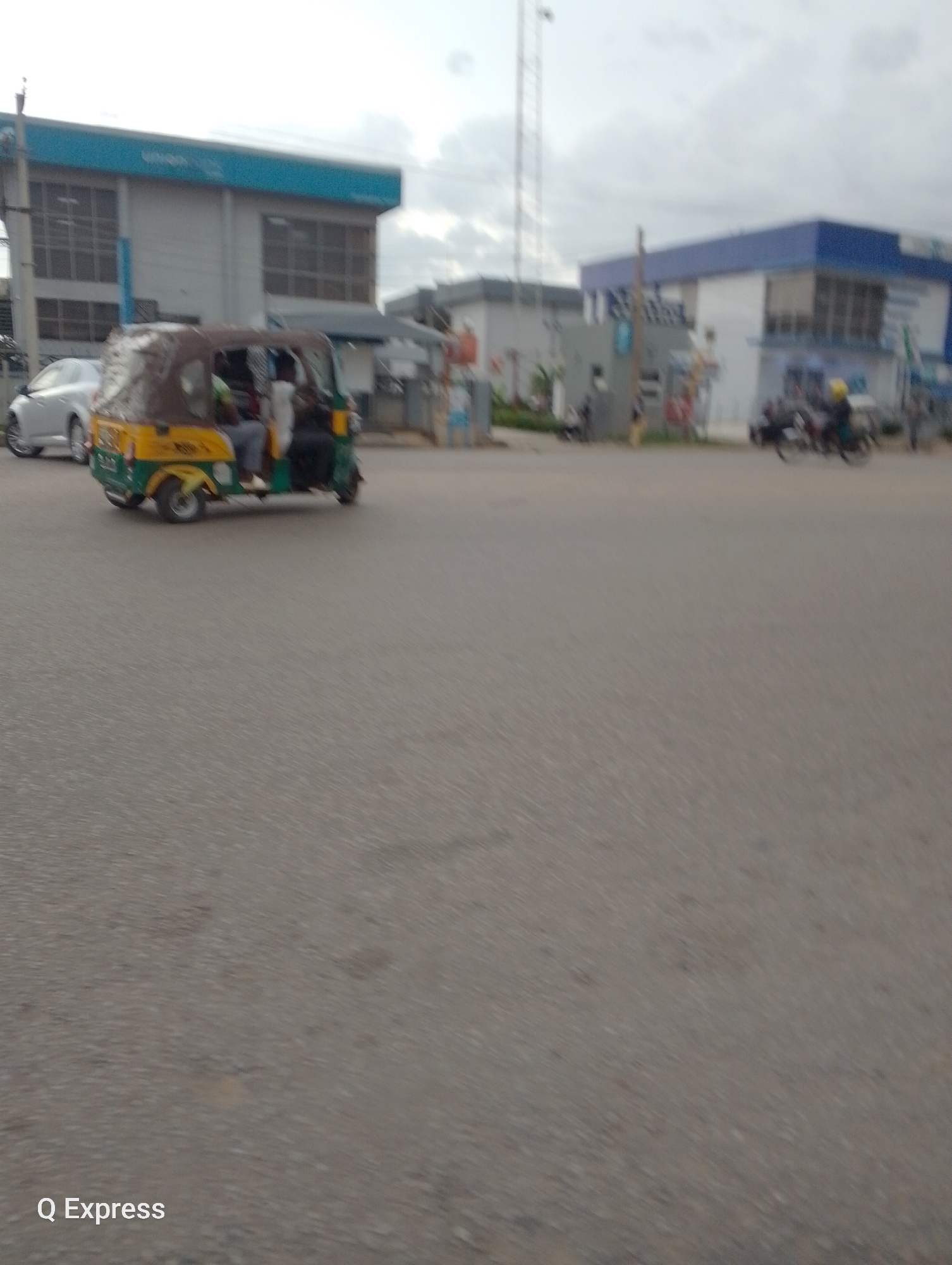 A street scene in Jos, Nigeria, features a motorized tricycle carrying passengers on a paved road. To the left, a silver car is parked. In the background, two buildings are visible: one with a blue stripe along the top and another with a prominent blue roof. A utility pole and what appears to be a communication tower stand in the distance. The sky is overcast. The image also contains a watermark in the lower left corner reading "Q Express."