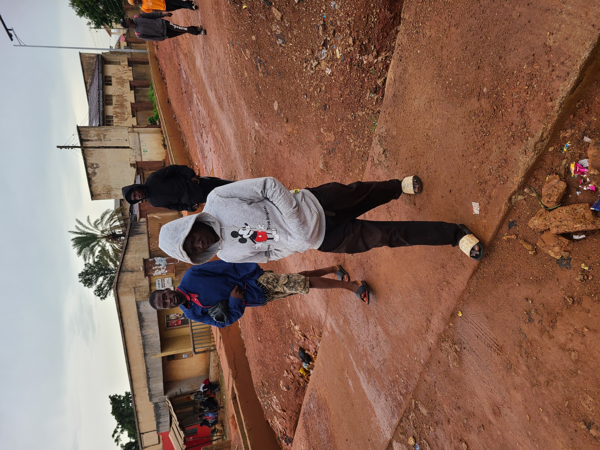 In Gura Top, Nigeria, five men walk along a muddy, red dirt road. The man in the foreground wears a grey hooded sweatshirt with a Mickey Mouse graphic and black pants. He has his hands in his pockets and is wearing patterned slide sandals. To his left, a man in a blue jacket and camouflage shorts walks with a black bag in his hand. He is also wearing slide sandals. Behind them, a man in a black hooded sweatshirt walks ahead. Further back, another man in a grey jacket with "Supreme" written on the back of his pants walks, followed by a man in an orange shirt. The background shows several buildings, some appearing to be shops or residences, with a cloudy sky above. The road is uneven and littered with debris.