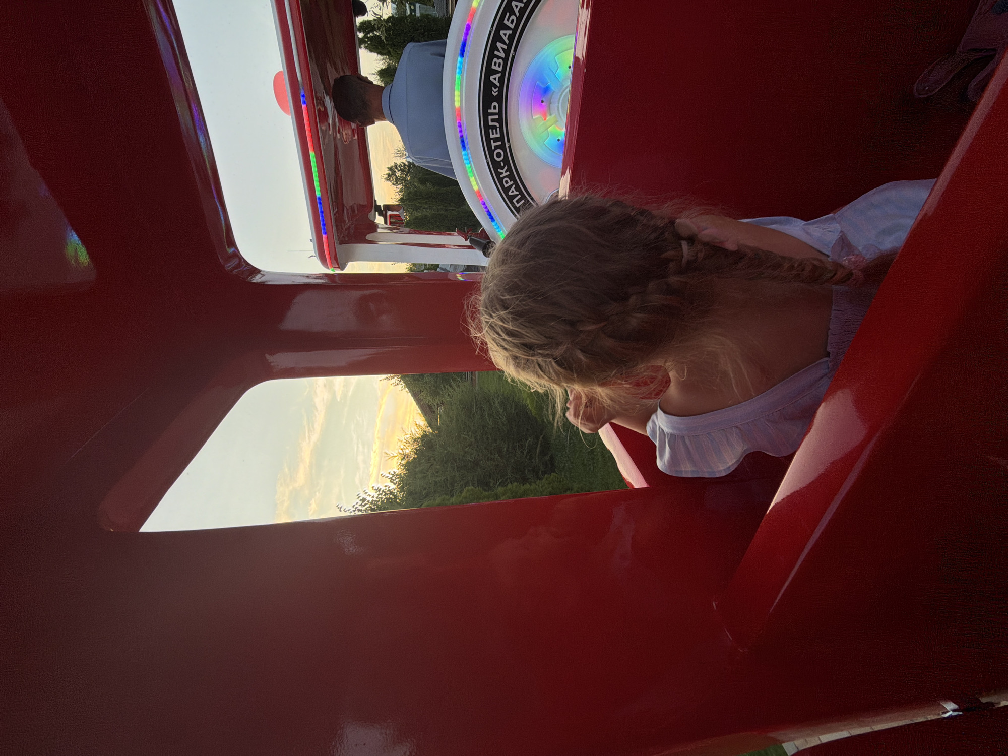 In Kamyshin, Russia, a young girl with blonde hair braided into a pigtail is seated inside a red amusement park train car, looking towards the setting sun. She is wearing a light-colored dress. In front of her, a man in a blue shirt is visible from the back, operating the train. The train is decorated with colorful LED lights, and a circular emblem with Cyrillic text reads "ПАРК-ОТЕЛЬ «АВИАБА»" (Park-Hotel "Aviaba"). The scene takes place outdoors, with greenery and trees visible in the background against a twilight sky.
