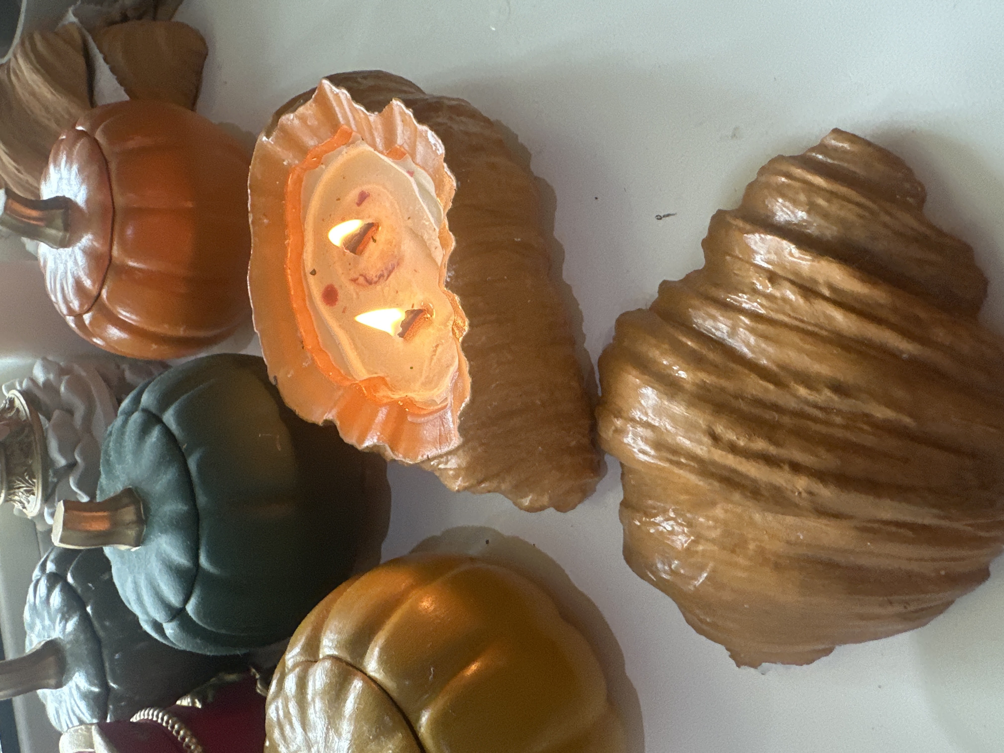 The image displays a collection of decorative, pumpkin-shaped ceramic containers and a croissant-shaped candle on a white surface. Some of the pumpkin containers are green, while others are orange. The croissant candle is lit, with two visible flames. The overall scene suggests a festive or autumnal decoration. There are no discernible elements that would indicate a specific location within Vladivostok, Russia.