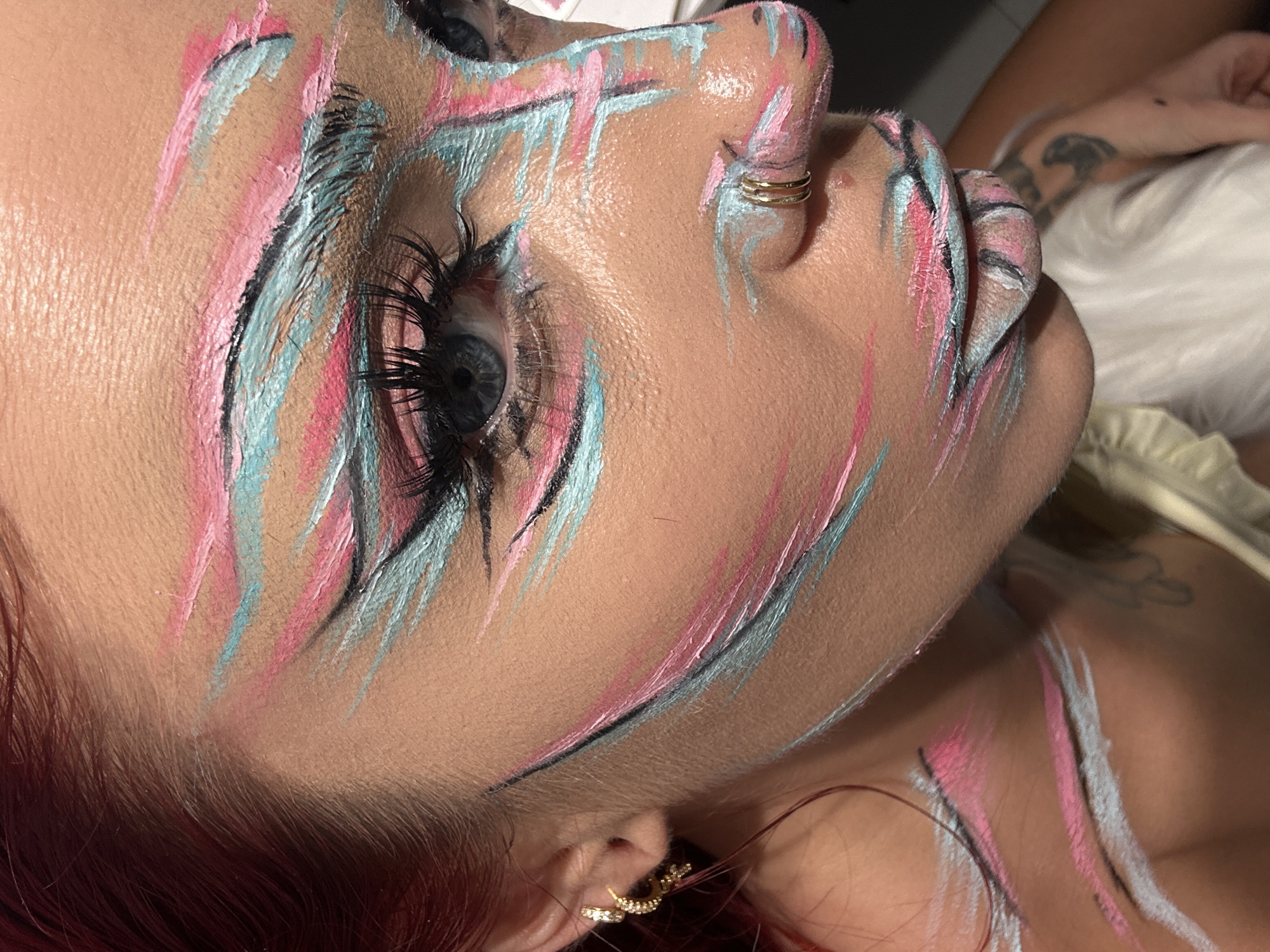 A close-up, profile view of a woman's face with abstract, colorful face paint. The paint features streaks of pink, light blue, and black, applied in a dynamic, brushstroke-like fashion across her forehead, eyebrows, cheeks, and lips. She has on false eyelashes and a gold hoop earring in her left ear, and a double gold hoop piercing on her right nostril. Her blue eyes are visible, looking to her left. The background is blurred and dark. The image does not provide any information about a location in North Kuta, Indonesia.