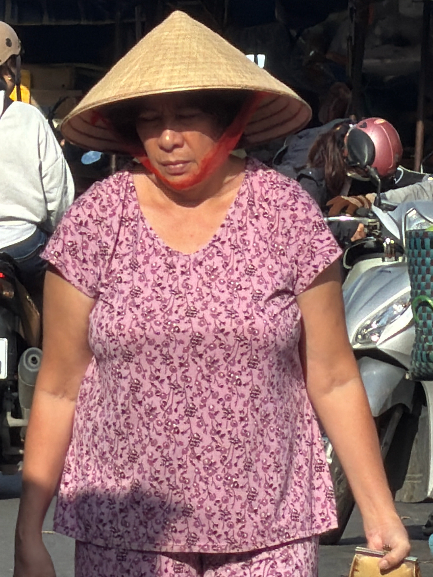 In Nha Trang, Vietnam, a woman wearing a conical hat and a pink floral shirt stands in what appears to be a market. She is holding a small, gold-colored coin purse. A motorcycle, complete with a red helmet, is visible behind her. The lighting suggests it is daytime, with shadows cast across the scene.