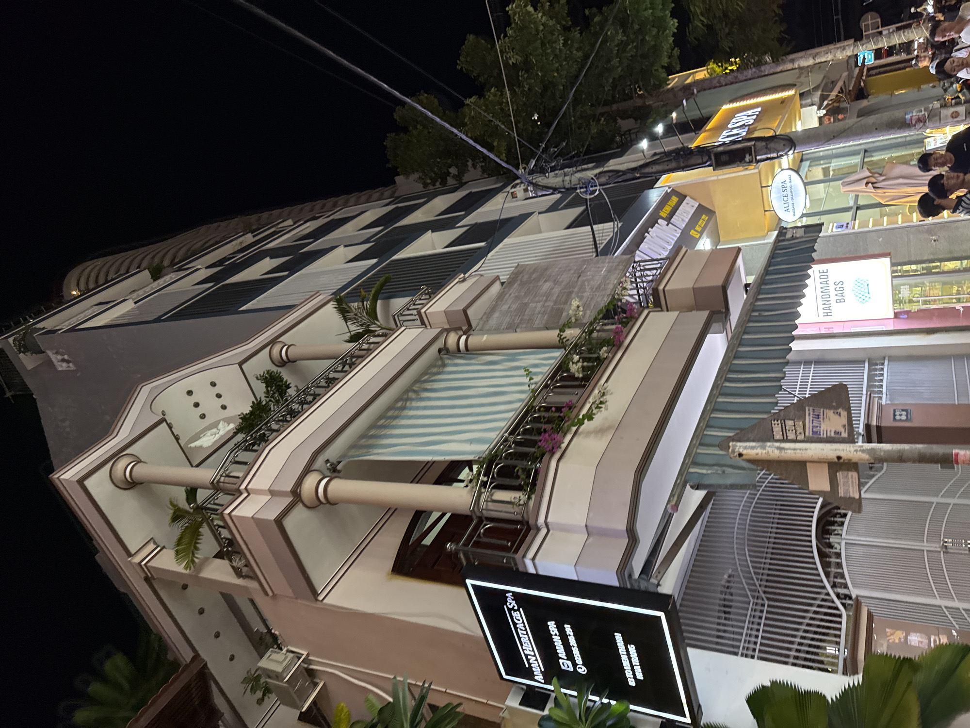 The image captures a nighttime street scene in Nha Trang, Vietnam. A multi-story building with a modern facade is illuminated, featuring balconies with railings and potted plants. A prominent sign on the left side of the building reads "AMAN HERITAGE SPA" and includes contact information and an address at "63 TO HIEN THANH, NHA TRANG." Below this, a large metal gate suggests an entrance to a property.
Further down the street, other illuminated signs are visible, including one for "ALICE SPA" which advertises "MASSAGE - SHAMPOO - NAILS." Another sign displays "HANDMADE BAGS." Several people are present on the sidewalk, some appearing to be customers or passersby. Electrical wires crisscross overhead, and trees line the street. The overall impression is of a commercial area in an urban setting at night.