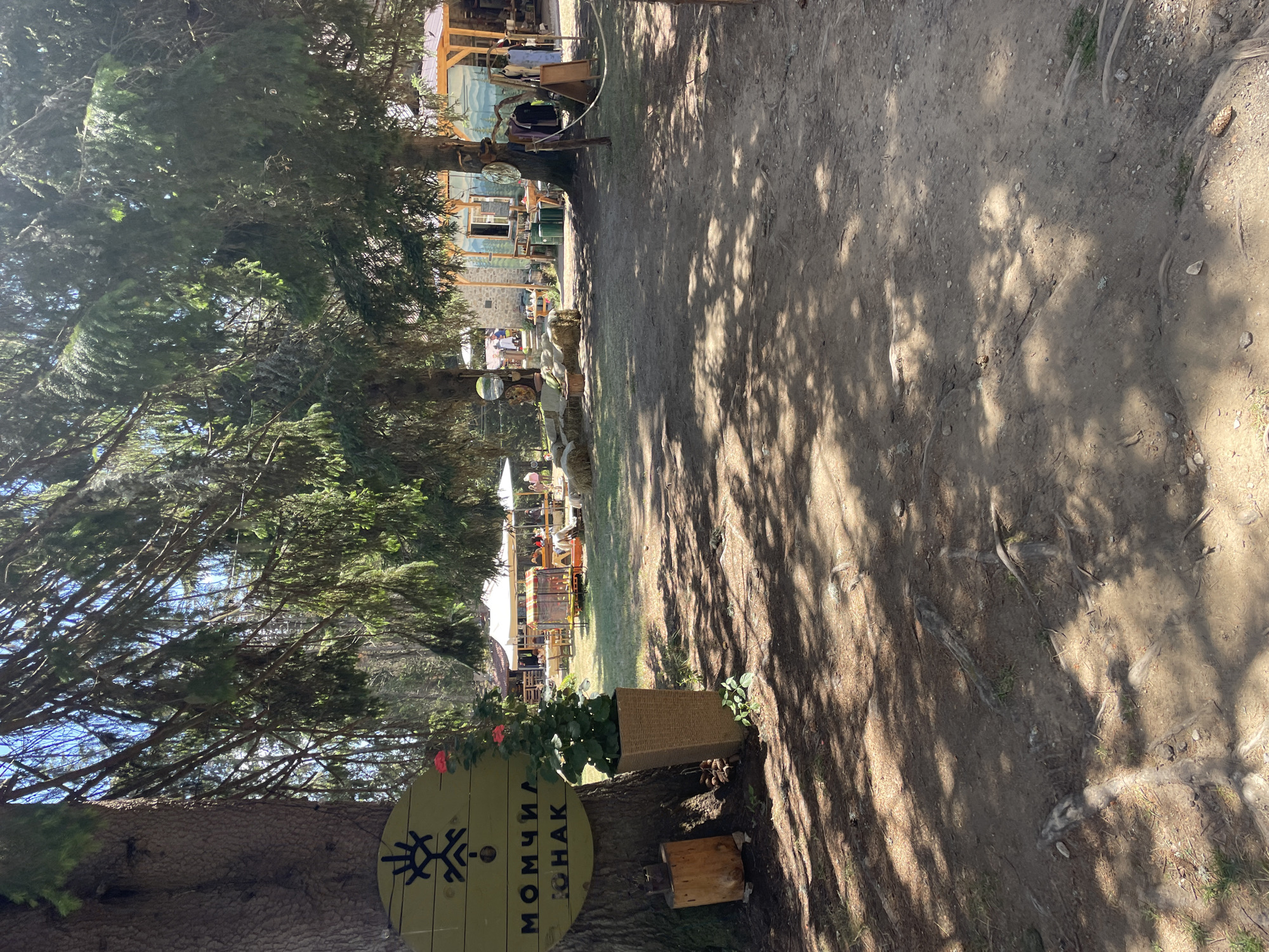 The image depicts an outdoor scene at a location identified as "Momchilova Laka" in Smolyan, Bulgaria. The foreground is a dirt path with exposed tree roots and scattered pinecones, dappled with sunlight and shadow from overhead trees. To the left, a large tree trunk is visible with a round, yellow sign attached to it, featuring a black symbol and Cyrillic text. A potted plant with red flowers stands next to the tree. In the background, a cluster of buildings with wooden roofs and stone walls can be seen. Several tables and chairs are set up on a grassy area in front of the buildings, with white canopies providing shade. People are present, engaged in various activities, with some individuals walking and others seated at the tables. The environment suggests a rural or natural setting, possibly a tourist attraction or a recreational area.