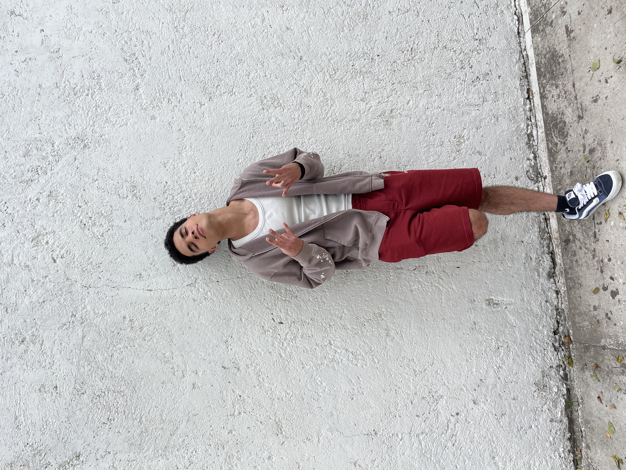 A young man is lying on a white textured surface that appears to be concrete, next to a concrete curb on the right. He is wearing red shorts, a white tank top, a taupe-colored zip-up sweatshirt. He is wearing black socks and a dark blue and white sneaker on his right foot. He is on his back, with his head pointing towards the top left of the image and his feet towards the bottom right. He is holding both hands up in a gesture. The location appears to be an outdoor, possibly urban, setting. The city or town is San Bernardino Tlaxcalancingo, Mexico.