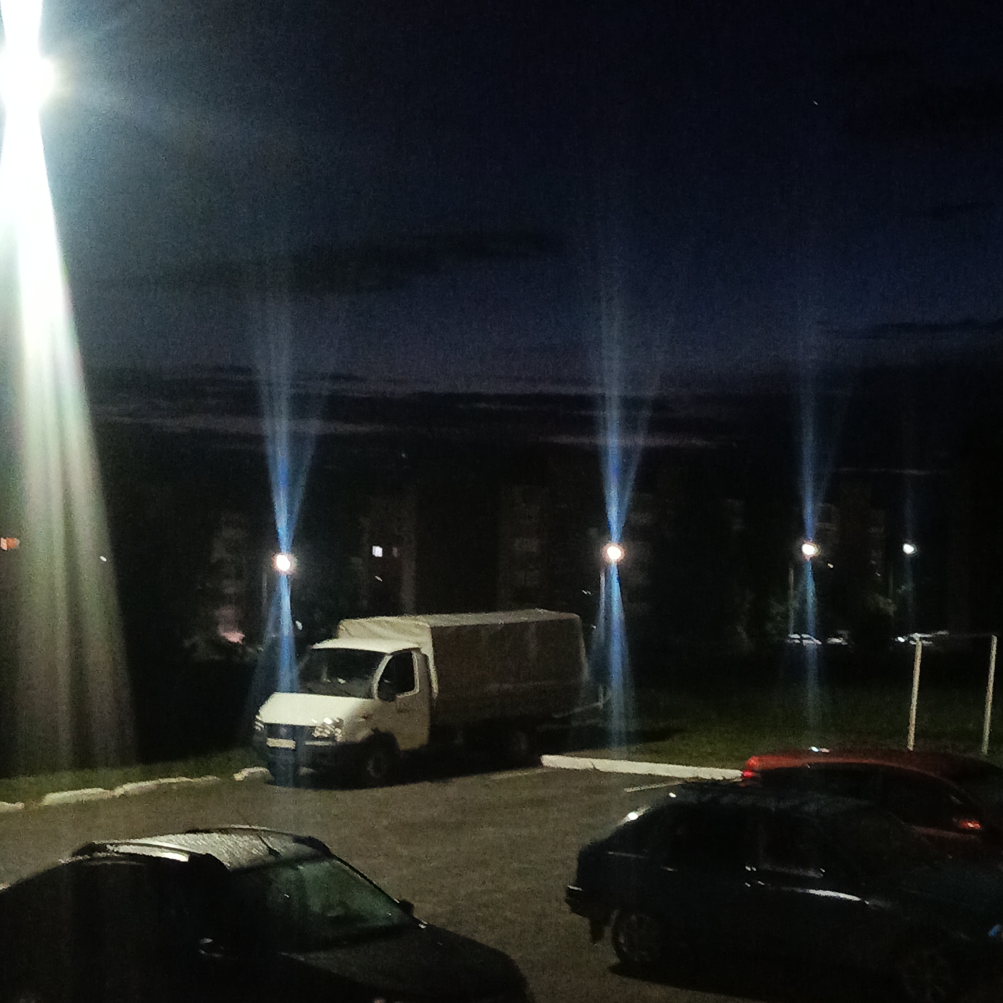 This image captures an outdoor night scene in Markovskiy, Russia, depicting a parking lot surrounded by buildings and foliage. The sky is dark, suggesting late evening or night.
The primary subjects are several vehicles. In the mid-ground, a white light commercial truck, likely a GAZelle, with a canvas-covered cargo bed, is parked prominently. In the foreground, a dark-colored car is partially visible on the bottom left, and another dark car is seen on the bottom right, with a red car parked directly behind it. The reflections of the night lights are visible on the wet or glossy surface of the dark car in the foreground.
The scene is dramatically lit by multiple artificial light sources. A very bright, overexposed light source on the far left casts a wide, intense beam of light. Several streetlights are scattered throughout the mid-ground and background, each producing distinct, blue-tinted vertical light streaks or lens flares, a common effect in night photography. Faint outlines of multi-story buildings are visible in the distant background, interspersed with dark masses of trees or bushes. Some small, distant lights suggest illuminated windows within these buildings. The ground consists of asphalt pavement for the parking lot and patches of green grass along its edges.
There are no people visible in the image, nor is there any discernible text. The overall atmosphere is quiet and still, characteristic of a residential or commercial area during nighttime.