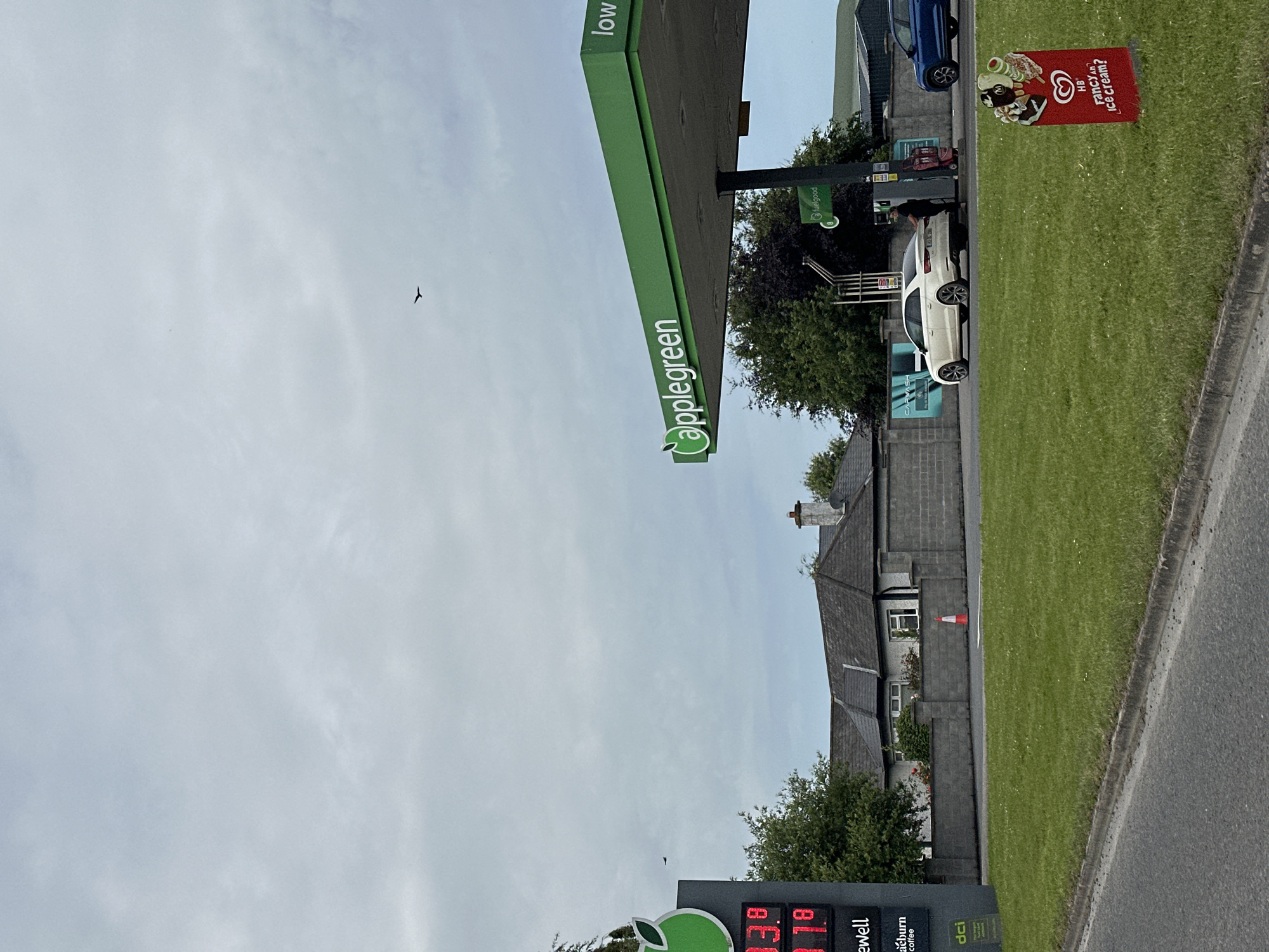 The image depicts an outdoor scene at an Applegreen gas station under an overcast sky. In the foreground, a section of a road and a grassy verge are visible. Further back, the gas station canopy with the "Applegreen" logo is prominent. To the left, a digital price board displays prices in red: "33.9" and "3.18". A white car is parked at a pump, with a person in dark clothing standing next to it. A blue car is also visible at another pump. A red sign advertising "HB Fancy an ice cream?" with images of ice cream is placed on the grass. Behind the gas station, a grey stone wall and a building with a dark roof are seen. In the sky, a bird is in flight. The scene appears to be in Ireland.