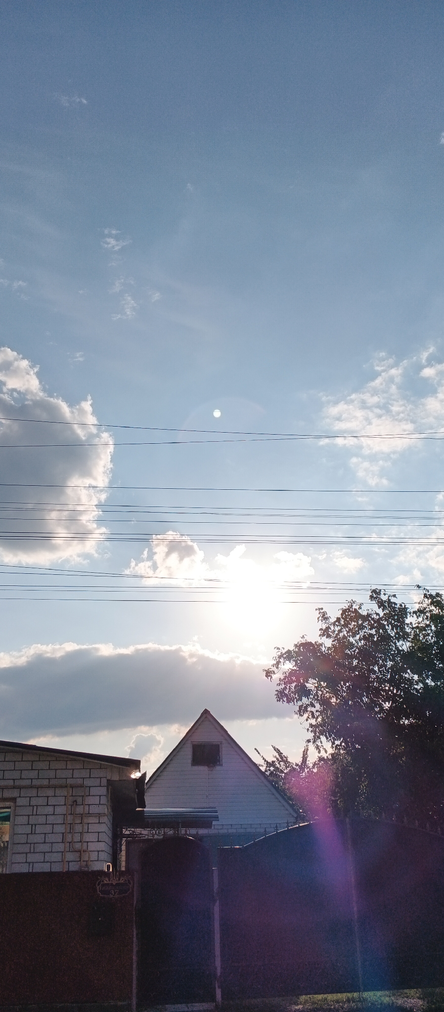 This is a vertical, eye-level outdoor photograph taken in Smila, Ukraine, depicting a bright, sunlit sky with scattered clouds and power lines crisscrossing the frame. In the foreground, a white brick building is partially visible on the left, and a gabled house with a dark-trimmed window is centered. A dark fence runs along the bottom of the image, with a tree on the right side casting a purple lens flare across the scene. The sun is bright and partially obscured by clouds, creating a lens flare in the upper center of the image.