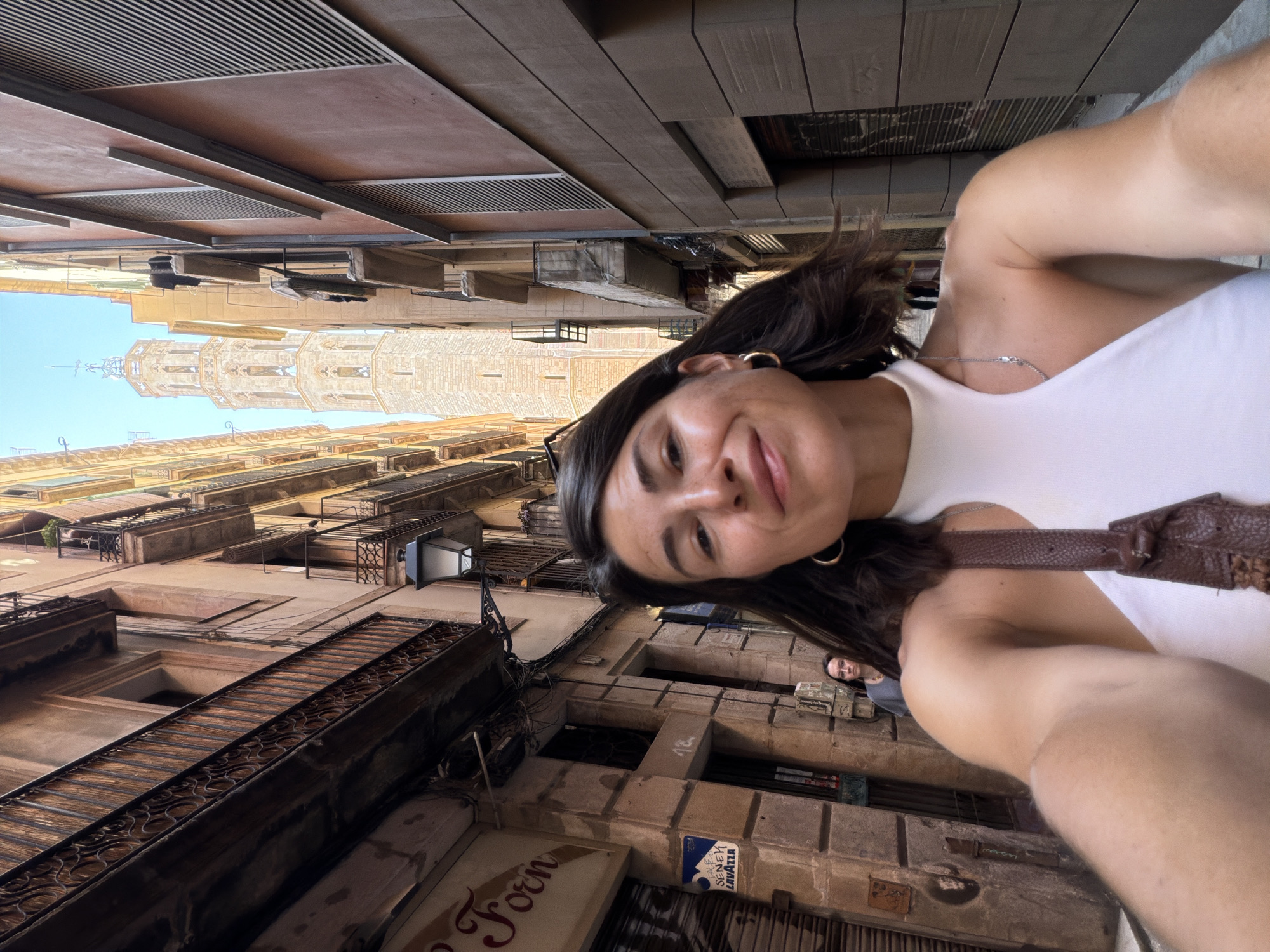 A woman is taking a selfie in a narrow European street with tall buildings on either side. A bell tower is visible in the distance. She is wearing a white halter top and a brown purse. The street is paved and there is a woman walking in the background. The buildings have balconies and shutters.