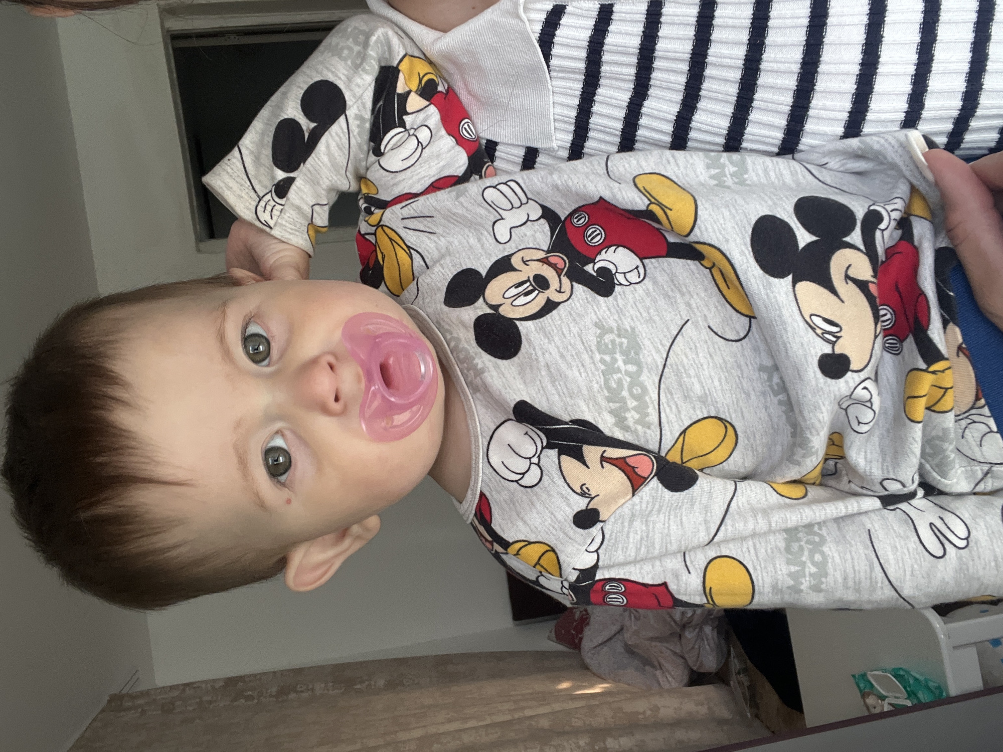 A close-up, eye-level medium shot shows a baby with green eyes and dark brown hair, wearing a gray Mickey Mouse pajama top and a pink pacifier. The baby is looking directly at the camera, with their right hand near their ear. A portion of a person's arm, wearing a black and white striped shirt, is visible on the right side of the frame. The background is blurred, showing a curtain on the left and a doorway on the right, suggesting an indoor setting. The scene appears to be in an apartment or house.