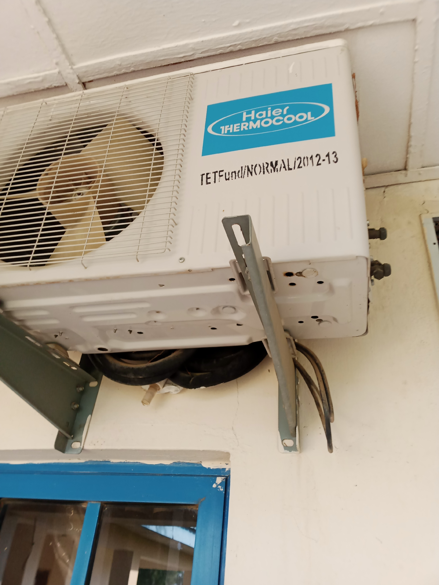 An outdoor unit of a Haier Thermocool air conditioner is mounted on a white wall beneath an overhang. The unit is white with a blue logo that says "Haier THERMOCOOL." Below the logo, text reads "TETFund/NORMAL/2012-13." The unit is supported by two metal brackets, one on the left and one on the right. The left bracket is partially visible and is gray. The right bracket is more prominent, also gray, and extends from the unit to the wall. Black electrical wires and hoses are bundled beneath the unit. Below the air conditioner, a blue-framed window with clear panes is visible. The wall appears to be slightly weathered, with some visible cracks and discoloration. The scene is set in Jalingo, Nigeria.