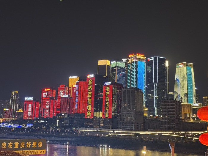 The image displays a nighttime panoramic view of the Yuzhong District skyline in Chongqing, China, observed across a body of water. Numerous modern high-rise buildings dominate the far riverbank, each illuminated. Several buildings on the left are predominantly lit in red, displaying Chinese characters such as "金桥百信律师所" (Jinqiao Baixin Law Firm) and "六号片" (Block Six). Other buildings exhibit lighting in golden yellow, blue, and white. A tall building on the far right features a large, multi-colored landscape projection depicting mountains and water in green and blue hues. An illuminated promenade with small, warm-toned lights runs along the river's edge. The river surface reflects portions of the city lights. In the lower-left foreground, a partially visible yellow sign displays the Chinese text "我在重庆好想你" and its Pinyin "CHONG QING HAO XIANG NI", accompanied by a right-pointing arrow. Two large, spherical, internally lit red Chinese lanterns are partially visible and out of focus in the lower-right foreground. The sky above is dark and clear, with a faint green light source visible in the upper center-left. No discernible individuals or direct human actions are present.