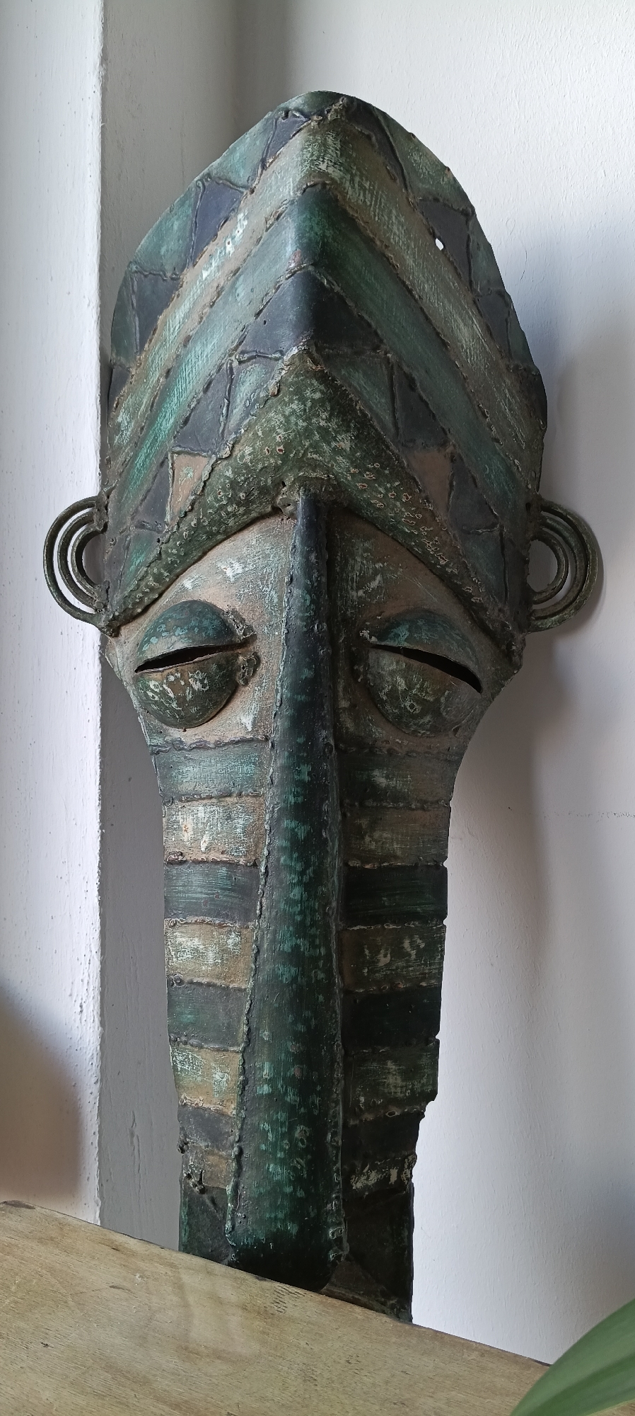 The image shows a close-up of an abstract, elongated mask with a metallic, weathered patina, predominantly in shades of green and bronze. The mask has a prominent, ridged nose, almond-shaped eyes with open slits, and coiled ear-like protrusions. The surface of the mask is textured and shows signs of age and wear, with visible seams and imperfections from its construction. It is positioned against a plain, textured white wall, with a portion of a light-colored wooden surface visible at the bottom of the frame. The lighting appears to be natural, casting subtle shadows and highlighting the mask's textured surface. There are no people, animals, or other identifiable objects in the scene. The mask's style suggests it could be an artifact of tribal or modern art. There is no visible information or context within the image to associate it with Linz, Austria.