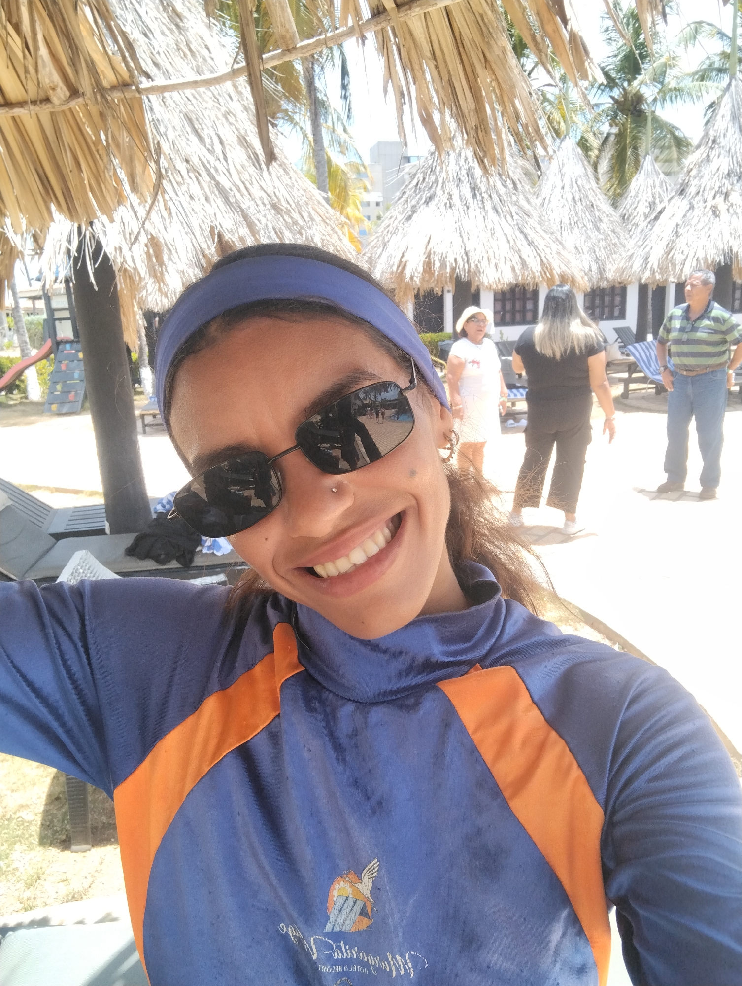 A woman wearing a blue and orange shirt, a blue headband, and sunglasses is smiling for a selfie. She is standing under thatched-roof structures, likely at a beach resort. In the background, other people are visible including a woman in white, a woman in black, and a man in a striped shirt and jeans. The location is likely Porlamar, Venezuela, based on the shirt's logo. Palm trees and a white building are also in the background.