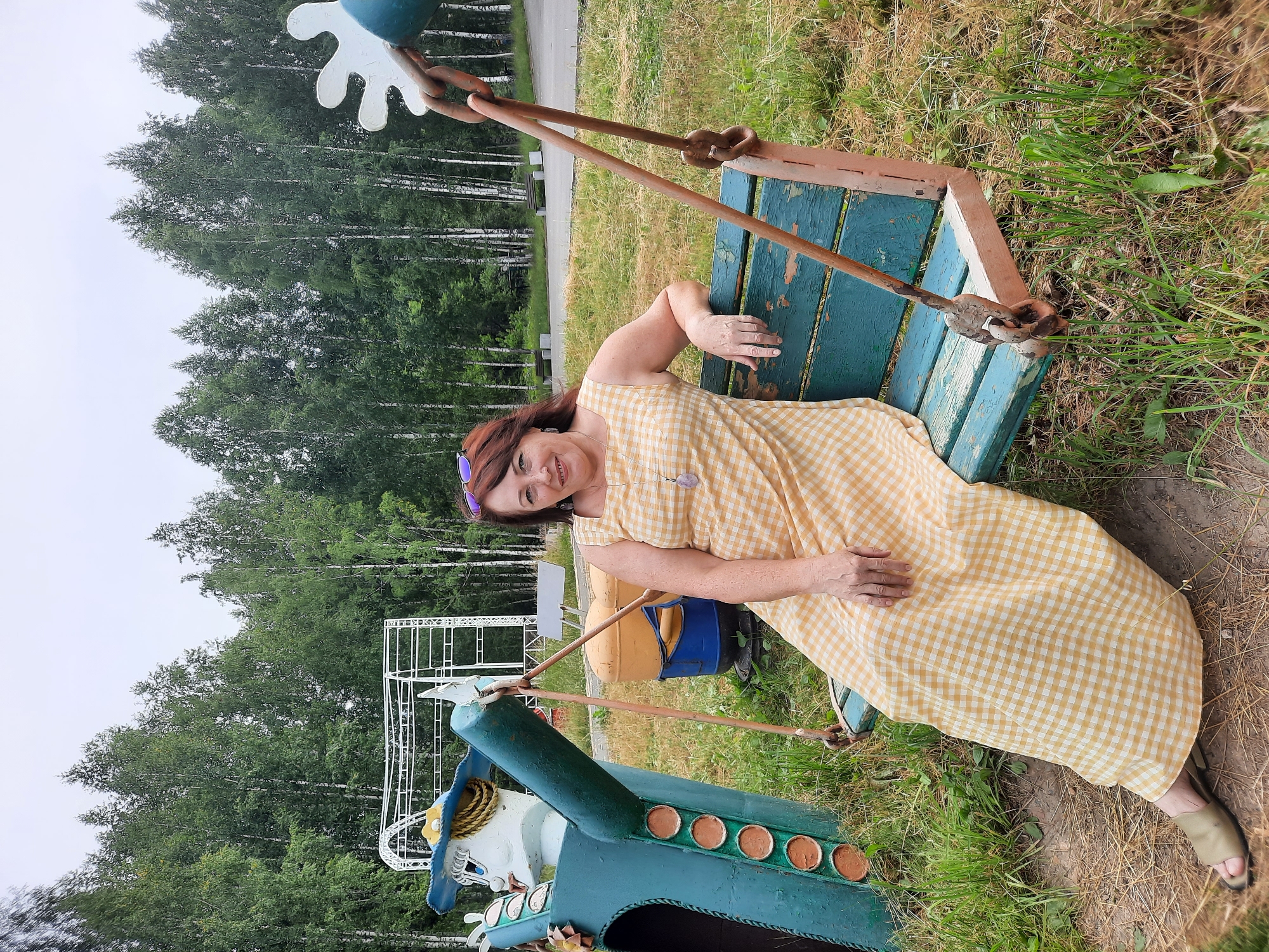 A woman with auburn hair, wearing a yellow and white checkered dress and gold sandals, is sitting on a weathered blue and brown swing. She is smiling at the camera, with her left arm resting on the back of the swing. Her right arm is bent, with her hand resting on her leg. She is wearing a necklace with a purple pendant and sunglasses are perched on her head. To her left, partially visible, is a large, teal-colored cartoon-like figure with a blue hat and yellow trim. The swing and figure are in a grassy area with scattered dry leaves. In the background, a line of birch trees forms a dense forest. A white metal structure, possibly part of a playground or carnival ride, is visible behind the cartoon figure. The scene appears to be outdoors in an amusement or park setting, possibly in the city of Zelenogorsky, Russia, given the prevalence of birch forests in the region.
