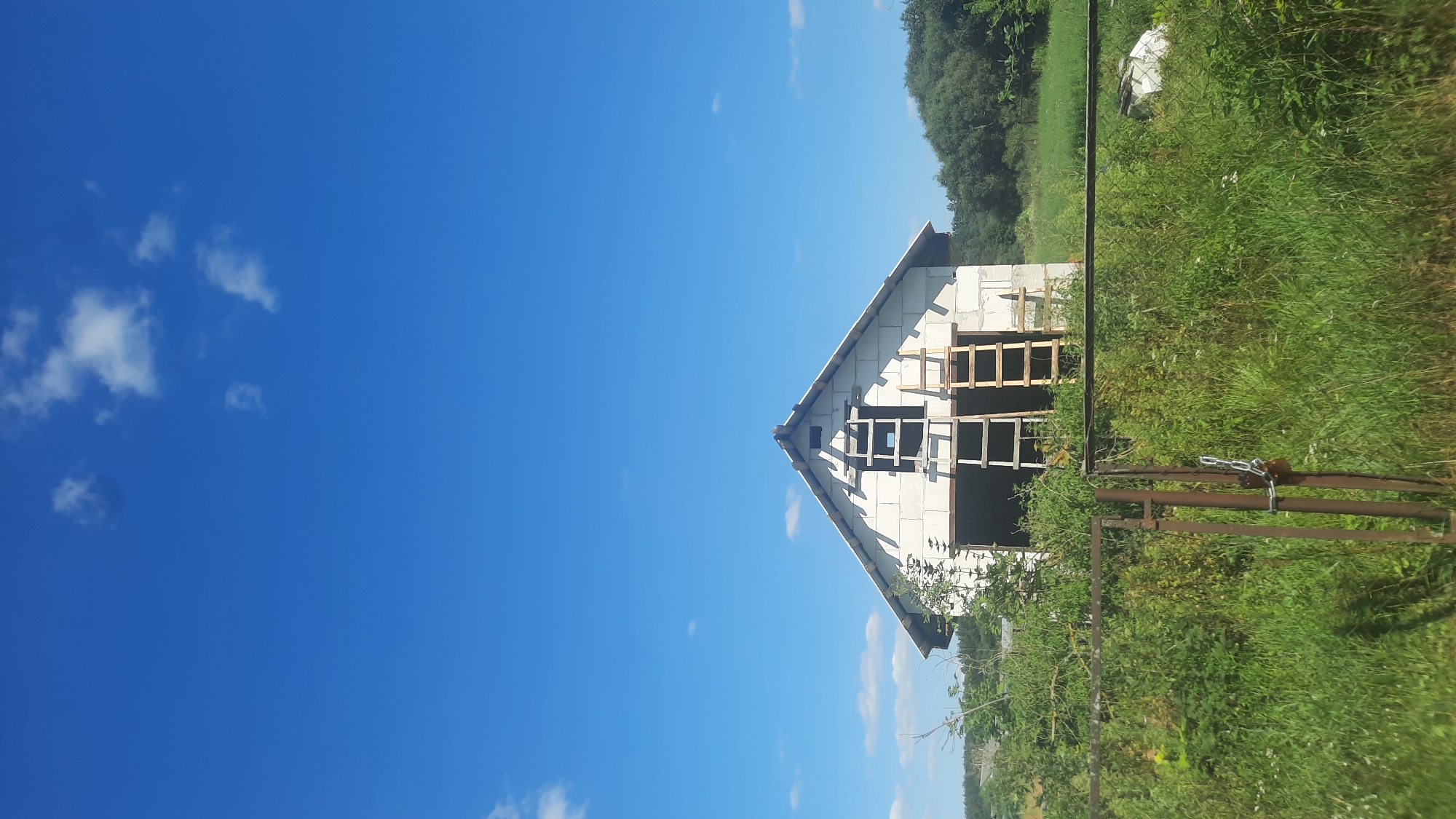 In the city of Shklov, Belarus, a partially constructed building with a white cinder block facade stands under a clear blue sky dotted with sparse white clouds. Two wooden ladders are propped against the building, suggesting ongoing construction or renovation. The building is surrounded by lush green vegetation, with tall grass and various plants growing around it. A rusty metal fence with a padlock secures the entrance to the property, further emphasizing the unfinished nature of the construction. In the background, a dense line of trees marks the edge of the landscape, creating a natural border for the scene. The overall impression is one of a rural setting where development is in progress.