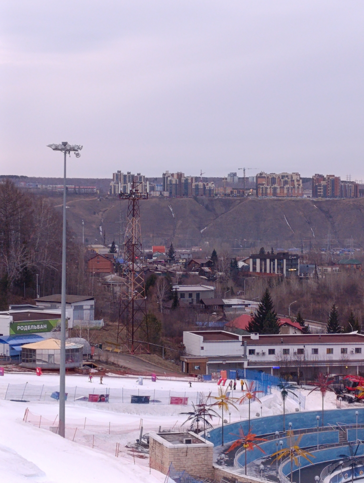 The image captures a wintry scene in Krasnoyarsk, Russia, featuring the Bobrovy Log Fun Park against a backdrop of the city's skyline.
Foreground: A light pole stands prominently on the left. A ski slope covered in snow is visible, enclosed by safety netting and featuring various advertising banners. The bottom right corner contains a decorative blue structure with artificial palm trees.
Midground: Buildings are interspersed with trees and other urban structures. A tall communication tower rises above the other buildings. Several people can be seen skiing or snowboarding on the slopes.
Background: The skyline of Krasnoyarsk stretches across the horizon, consisting of modern apartment buildings clustered on top of a hill. The sky is overcast, indicating a dull day.
Overall: The image depicts an urban landscape with a leisure park designed for winter sports, set against the backdrop of a modern city on a cold day.