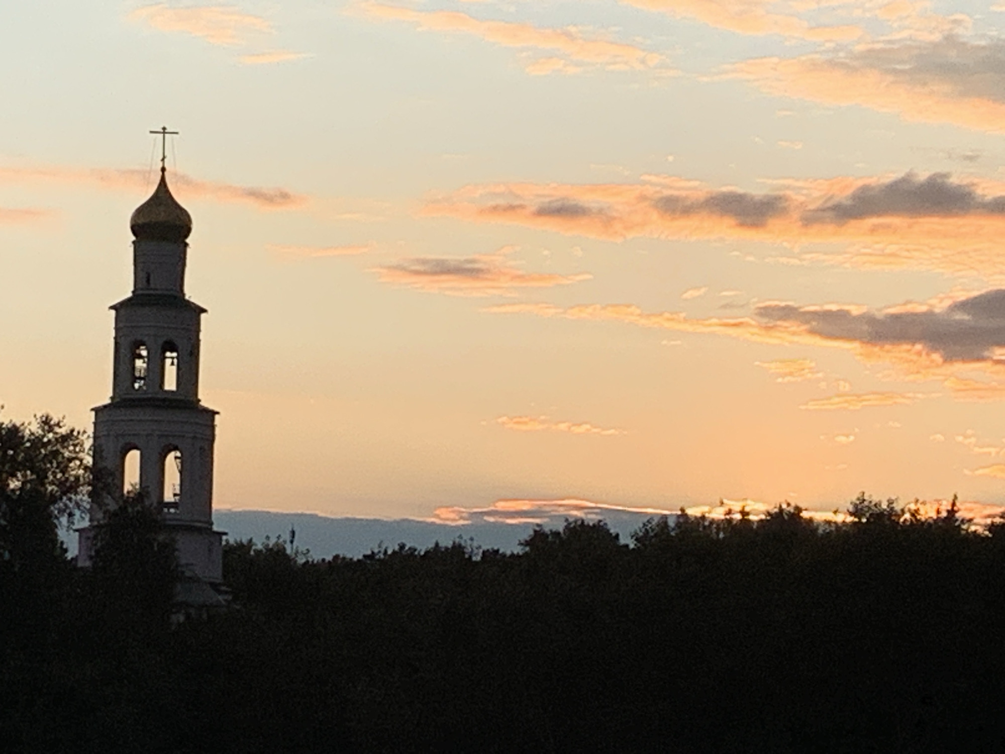 The image depicts a bell tower silhouetted against a sunset sky. The tower is light-colored with a golden dome topped by a cross. Arched openings are visible on the upper levels of the tower. In the foreground, a dense, dark band of trees stretches across the bottom of the image, obscuring the ground. The sky is a gradient of pale orange and yellow, with streaks of pink and gray clouds. The overall scene is peaceful and serene.
While the image itself does not provide specific information about the city of Perm, Russia, the architectural style of the bell tower, particularly the golden dome and cross, is characteristic of Orthodox churches found in Russia. Without additional context or landmarks within the image, it is impossible to definitively pinpoint the location within Perm.