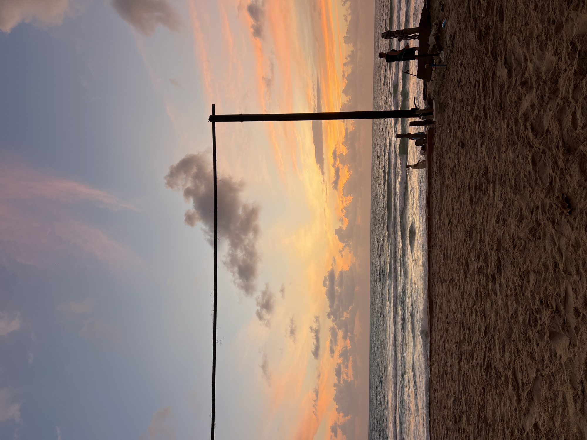This image depicts a beach scene at sunset in Choeng Thale, Thailand. A sandy beach occupies the foreground, with the ocean and a colorful sky filling the background. In the middle ground, several people are visible near the water's edge, appearing to be engaged in various activities, possibly fishing or setting up equipment. A large, dark, horizontal bar supported by a vertical post dominates the right side of the frame, extending across the upper portion of the image. The sky is a gradient of soft blues, pinks, and oranges, indicative of twilight, with scattered clouds. The ocean exhibits gentle waves rolling towards the shore. The overall impression is one of a tranquil evening on a tropical beach.