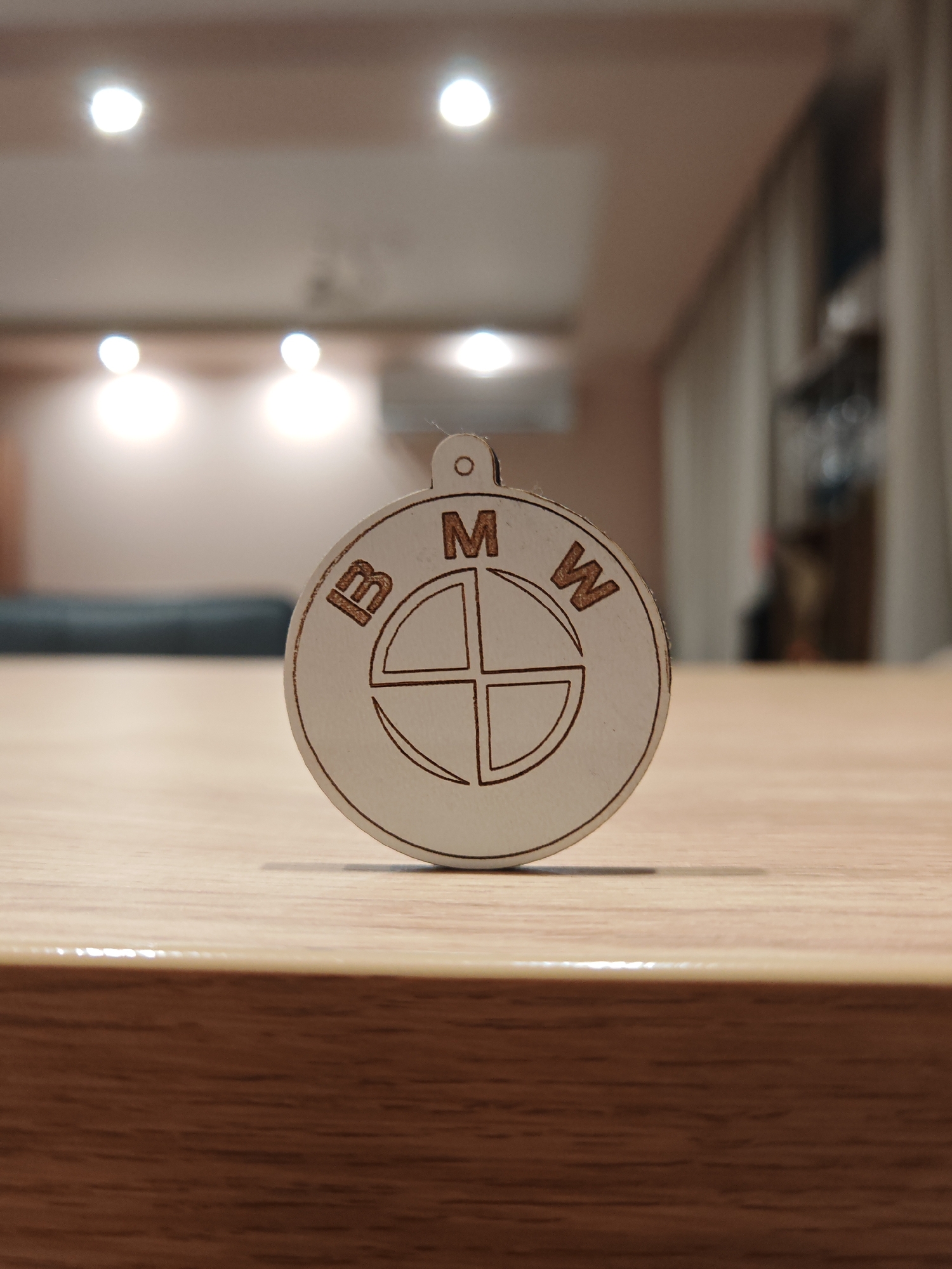 A close-up, eye-level shot captures a wooden keychain with the BMW logo. The keychain is circular, with the iconic blue and white circular emblem and the letters "BMW" etched into the wood. It is positioned on a light-colored, wooden surface, possibly a table. The background is blurred, showing a dimly lit room with ceiling lights and curtains in the distance. There are no people or animals visible in the image. The scene appears to be indoors, in a residential or office setting. The image does not contain any information that would allow for its location to be determined within the city of Plovdiv, Bulgaria.