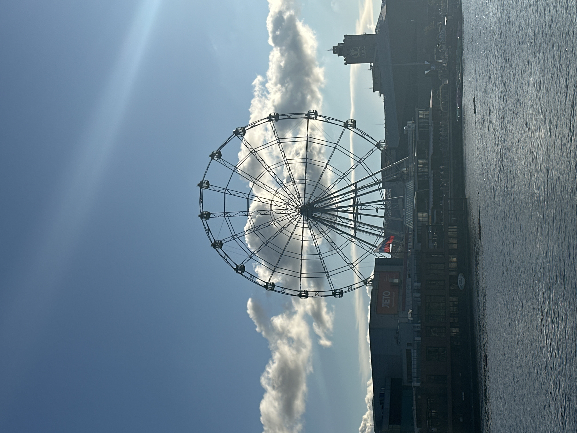A Ferris wheel dominates the center of the frame, its intricate metal spokes silhouetted against a bright blue sky dotted with scattered white clouds. The Ferris wheel appears to be in motion, or at least stationary with its cabins visible. In the foreground, a dark body of water reflects the light. Behind the Ferris wheel, a low-lying cityscape is visible, characterized by modern buildings and a prominent tower with a clock face. A Russian flag is visible on one of the buildings. The overall scene suggests an urban waterfront setting, possibly a park or amusement area.
