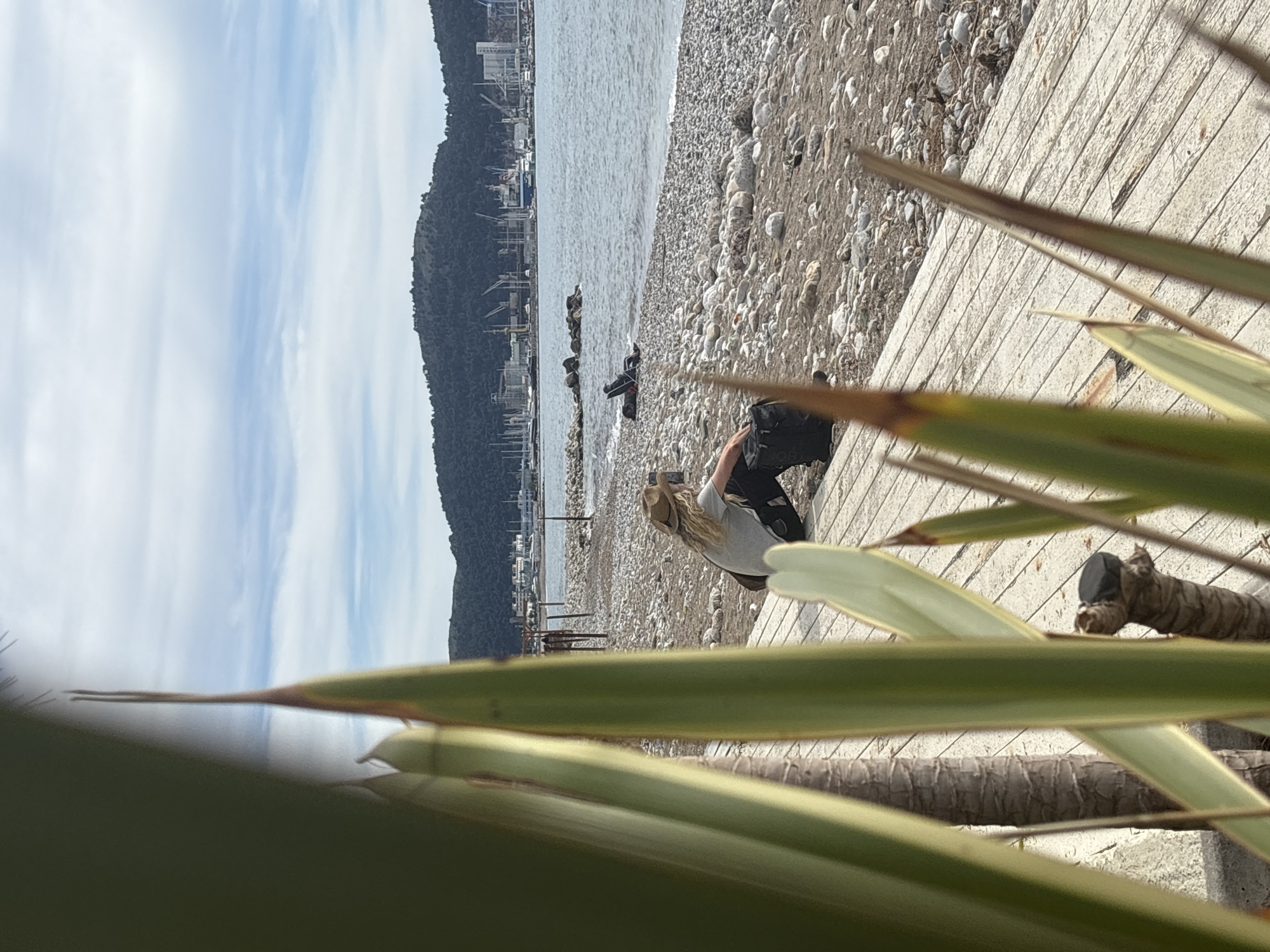 The image shows a seaside scene, likely a beach in Bar, Montenegro. The beach is made of pebbles and stones, with the Adriatic Sea stretching towards a backdrop of a mountain and the city of Bar in the distance. The sky is partly cloudy. In the foreground, there are green plants obscuring part of the view. A woman with blonde hair wearing a light grey shirt and dark bottoms stands near a wooden boardwalk, possibly interacting with some luggage or equipment. There are several dark objects situated at the water's edge which may be camping equipment, as well as some rusted construction objects.