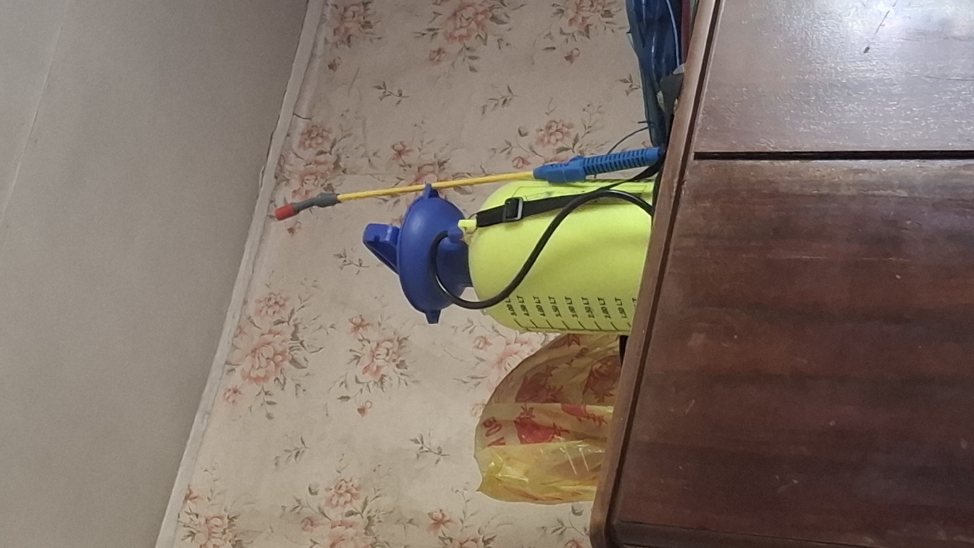 The image shows a close-up of a wooden cabinet top in Шұбарсу, Kazakhstan. On the cabinet top, there is a yellow plastic spray bottle with a blue lid and a yellow wand. Next to the spray bottle is a crumpled yellow plastic bag. The background consists of floral wallpaper. The lighting is dim, suggesting it is indoors.