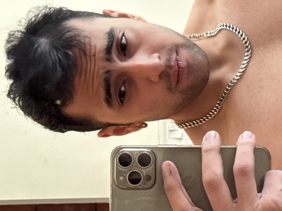 A shirtless man with dark hair and a gold chain around his neck takes a selfie in a mirror with a smartphone. The phone partially obscures his face, revealing his eyes, nose, and mouth, which is slightly upturned in a playful expression. The background appears to be a plain wall, with a door frame and light switch visible. The lighting is soft, creating shadows that highlight his features.