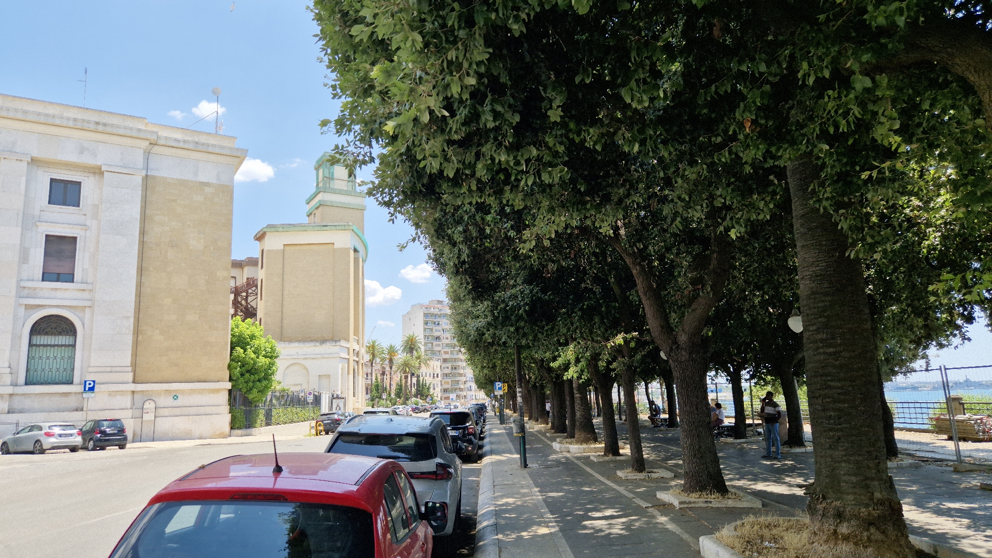 The image depicts a street scene in Taranto, Italy, on a sunny day. The foreground is dominated by the rear of a red car. To the left, a street runs alongside several parked cars, including a silver hatchback and a black mini-SUV. Behind them, a large, light-colored building with classical architectural elements stands prominently. Further down the street, other buildings of varying heights and styles are visible, including a taller, modern structure with many windows and a tower-like element on top.
A row of mature trees with dense green foliage lines the right side of the street, casting shadows on the sidewalk. Several people are visible on the sidewalk. One man in a blue shirt and jeans stands near a tree. Further down, other individuals are seated on benches under the trees. Beyond the trees, a body of water, likely the sea, is visible, with a fence and construction materials along the waterfront. The sky is blue with scattered white clouds. The overall impression is a city street with a mix of historical and modern architecture, lined with trees and bordering a waterfront.