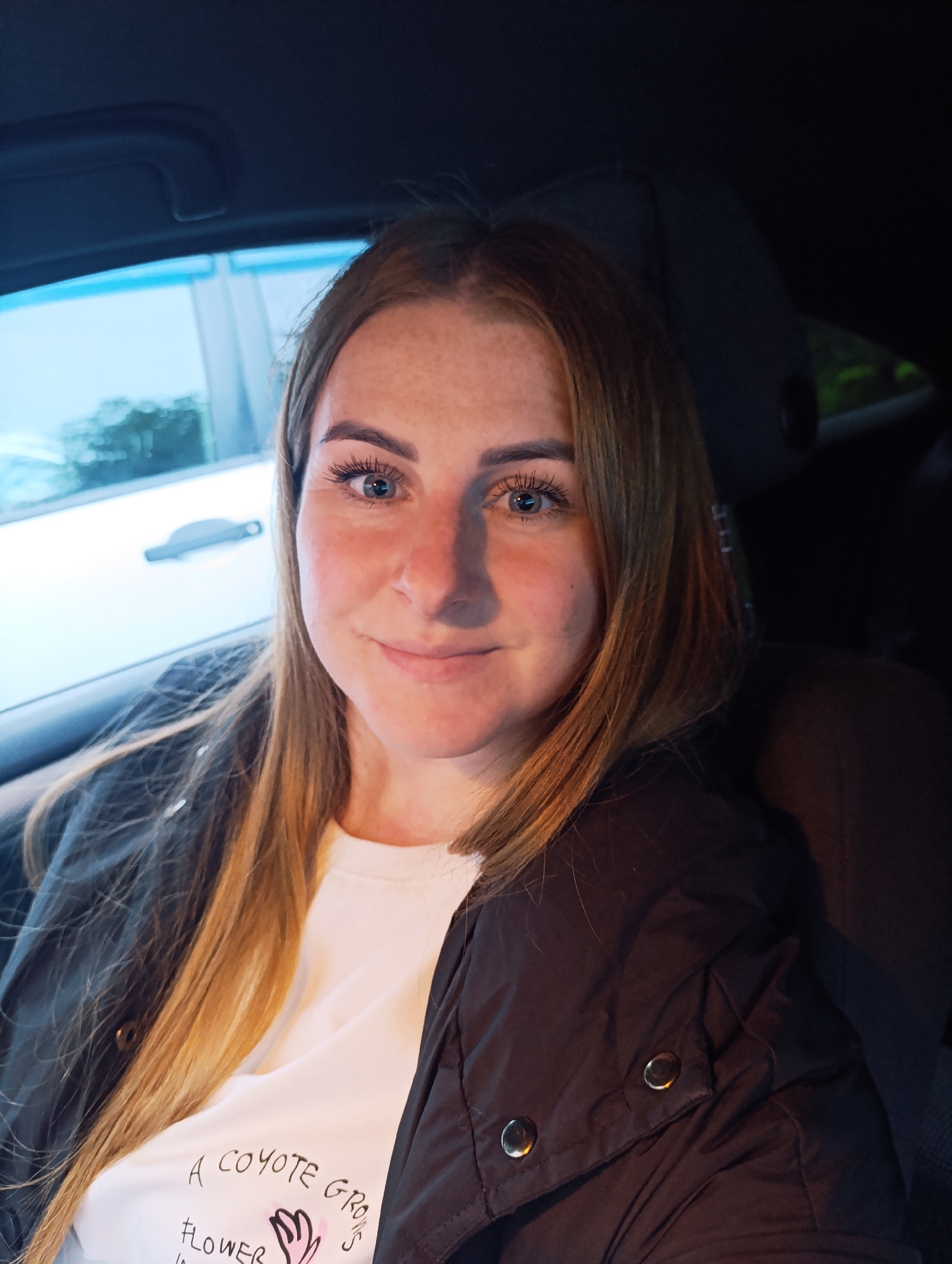 A young woman with long blonde hair and blue eyes is seated in the passenger seat of a car in Unknown, Unknown. She is wearing a white t-shirt with writing on it and a dark jacket. The car window shows a view of another car. The interior of the car is dark, with the seat and headrest visible.