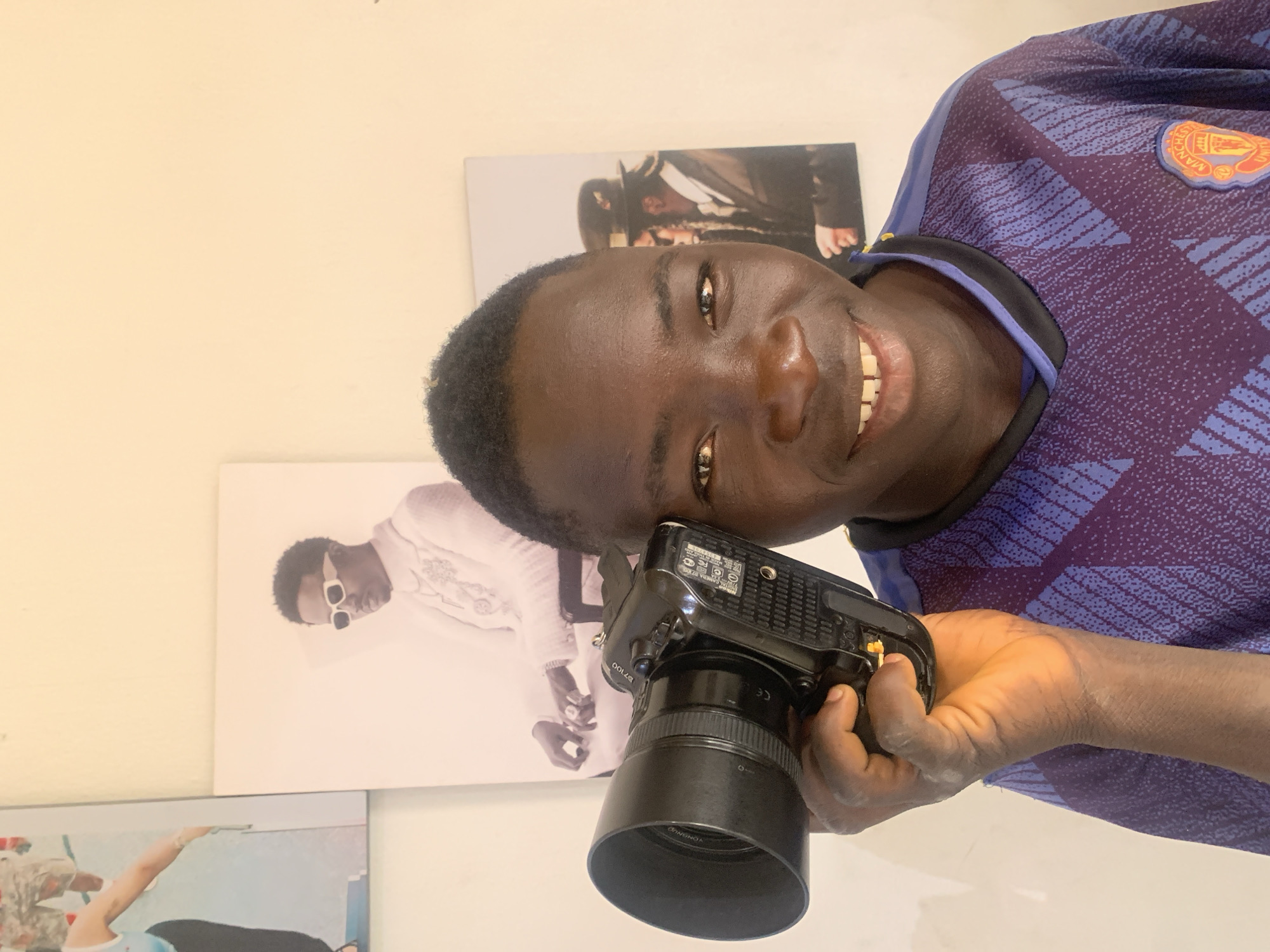 A vibrant portrait captures a young Black man in an indoor setting in Jos, Nigeria, smiling broadly and looking directly at the viewer. He wears a dark blue and purple patterned long-sleeved shirt, which appears to be a Manchester United football jersey, indicated by the partially visible team emblem on his right shoulder. In his left hand, he holds a black DSLR camera, identified as a Nikon, likely a D7100 model, with a prominent black lens and a "YONGNUO" branded lens hood. Visible text on the camera includes "NIKON CAMERA" and "MADE IN CHINA."
The background consists of light-colored walls decorated with several posters. One poster behind his head features a person in a dark suit. Another, below and to his left, depicts a figure resembling musician Wizkid, dressed in a white outfit and sunglasses. A third poster is partially visible at the bottom left. The man's cheerful expression and his grip on the camera suggest an enthusiasm for photography, possibly portraying him as a photographer. The lighting indicates it is daytime.