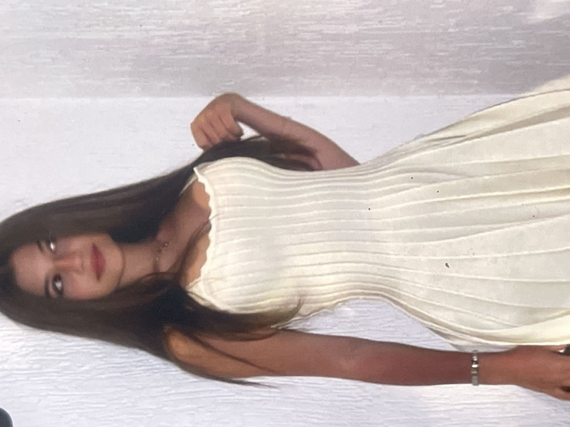This image captures a young woman lying down in an indoor setting, likely in Poltava, Ukraine, given the provided context. The photograph is oriented horizontally, showing the woman's body extending across the frame from left to right.
The primary subject is a young woman with long, dark brown hair that cascades over her shoulders and extends onto the white surface beneath her. She has a calm expression, with light-colored eyes looking towards the camera and her lips colored with red lipstick, suggesting a slight smile. She is wearing a light-colored, possibly off-white or cream, ribbed top or dress with thin straps, which accentuates her figure. A delicate necklace with a small pendant is visible around her neck, and a silver-toned bracelet, possibly adorned with small red or dark details, is on her right wrist.
The woman is posing while lying down, with her left arm bent and her hand resting near her head, though mostly obscured by her hair. Her right arm extends down the frame. The background is a plain, light-colored (white or off-white) textured surface, possibly a bed, blanket, or soft flooring, which creates a clean and simple aesthetic.
The time of day and weather are not discernible from the indoor setting. The lighting appears soft and even, highlighting her features without harsh shadows. There is no visible text in the image. The overall mood is serene and composed.