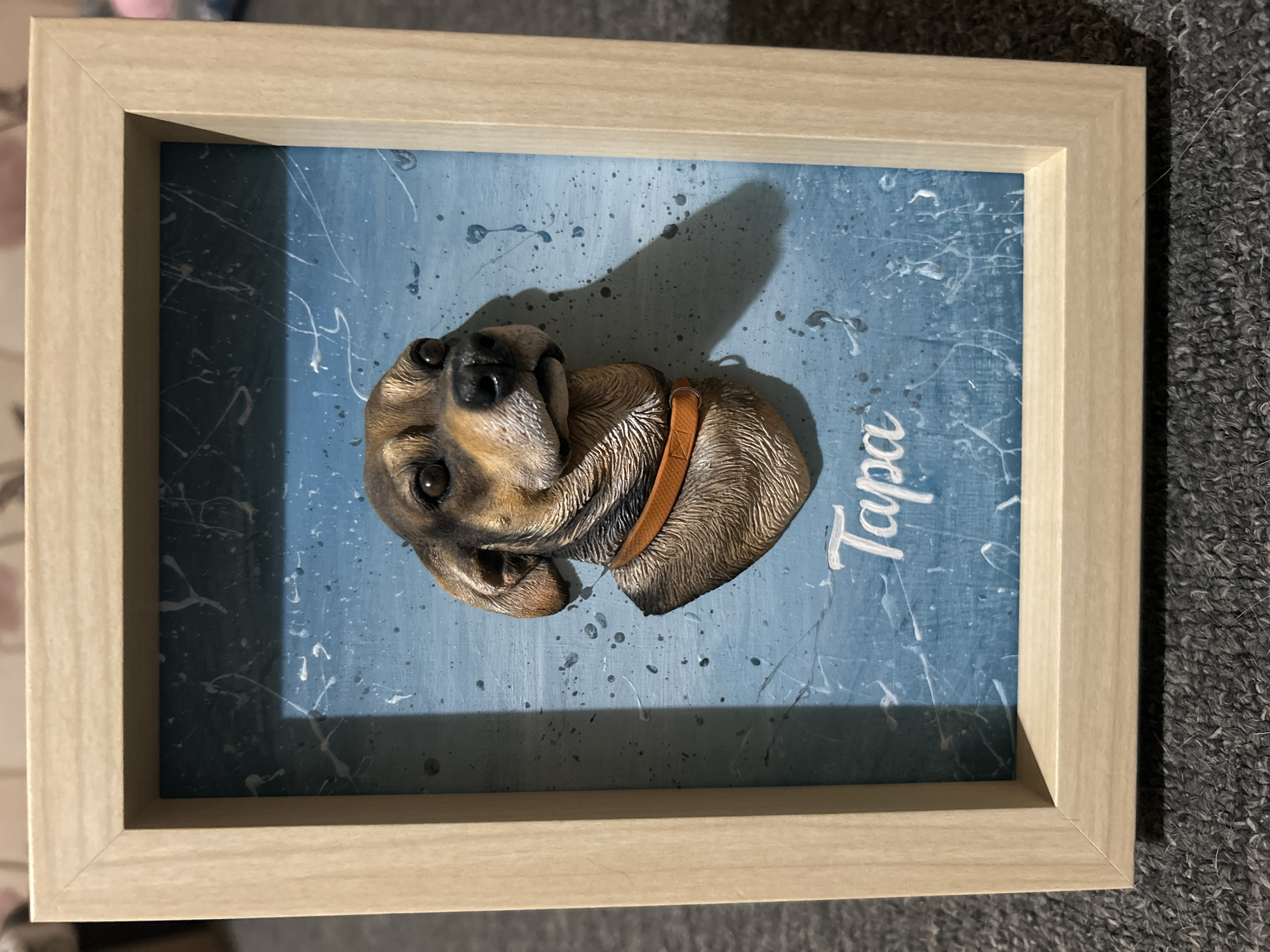 A framed, three-dimensional sculpture of a dog's head is centered in the frame. The sculpture is detailed, showing the dog's fur texture, eyes, nose, and ears, and features a tan collar. The dog's name, "Tapa," is written in white cursive script below the sculpture. The background is a textured blue surface with white paint splatters and drips, creating a distressed or abstract effect. The frame is made of light-colored wood. The image is taken from a slightly elevated angle, with the artwork resting on a dark gray, carpeted surface.