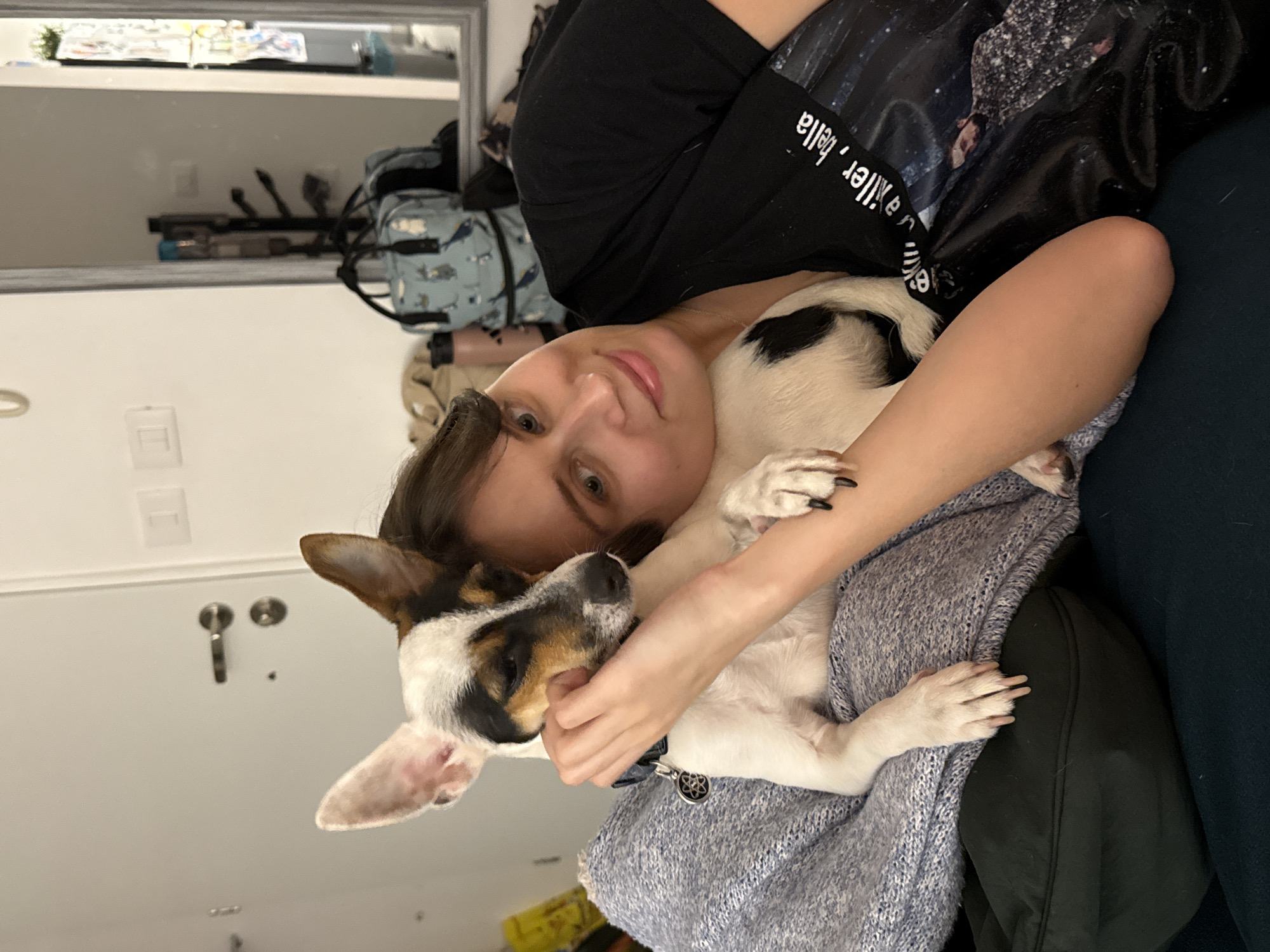 A young woman is lying on a dark blue couch, embracing a small, white and brown dog. She is wearing a black t-shirt with white writing on the sleeve. The dog has black nails and is wearing a collar with a medallion. The woman is holding the dog close to her chest. A light blue knit blanket is visible beneath them. In the background, there is a mirror reflecting a cluttered shelf and a backpack. The location is not discernible from the image and cannot be determined as being in Tel-Aviv, Israel.