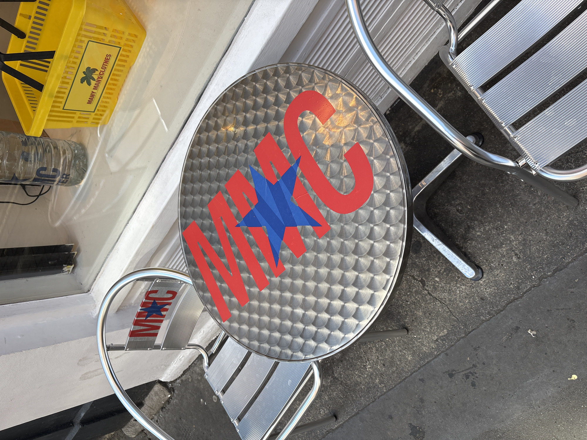 Captured from above, the image showcases a round metal table emblazoned with a logo featuring a blue star overlaid with red lettering. Two metal chairs with horizontal silver slats are positioned around the table. To the side, visible through a window, sits a yellow plastic basket with the words "MANY MANY CLOTHES" printed on it. The location is indicated by the branding on the table and chair as being affiliated with the "MANC" clothing brand in London, United Kingdom. The scene implies an outdoor seating area, possibly of a cafe or shop. The ground is paved with asphalt.