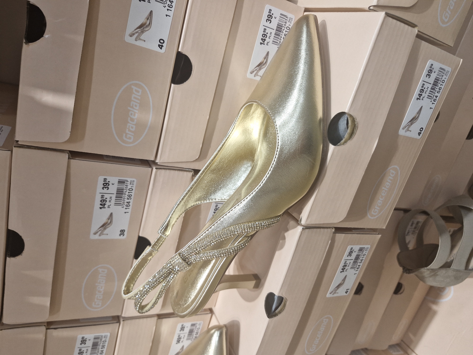 The image displays a close-up view of a metallic gold high-heeled shoe, likely a slingback pump, resting on top of stacked light brown cardboard shoe boxes. The shoe has a pointed toe and a medium-height heel, with its slingback strap intricately adorned with multiple rows of sparkling rhinestones. The interior of the shoe is also a bright gold color.
Numerous shoe boxes, primarily in shades of light brown or beige, are visible in the foreground, mid-ground, and background. Each box prominently features the brand name "Graceland" enclosed within a white oval logo. Many boxes also have price labels attached, consistently showing "149.99 PL PLN" and "39.99 €". These labels include barcodes, small illustrative drawings of a high-heeled shoe, and size indications such as "38", "40", and "41". Additional product identification numbers, like "1 164 56101" (with variations) and longer manufacturer codes (e.g., "4069919426819", "4069919426833"), are also printed on the labels. The scene suggests a retail environment or a shoe storage area, with the boxes neatly stacked. The lighting appears to be artificial, typical of an indoor commercial setting.