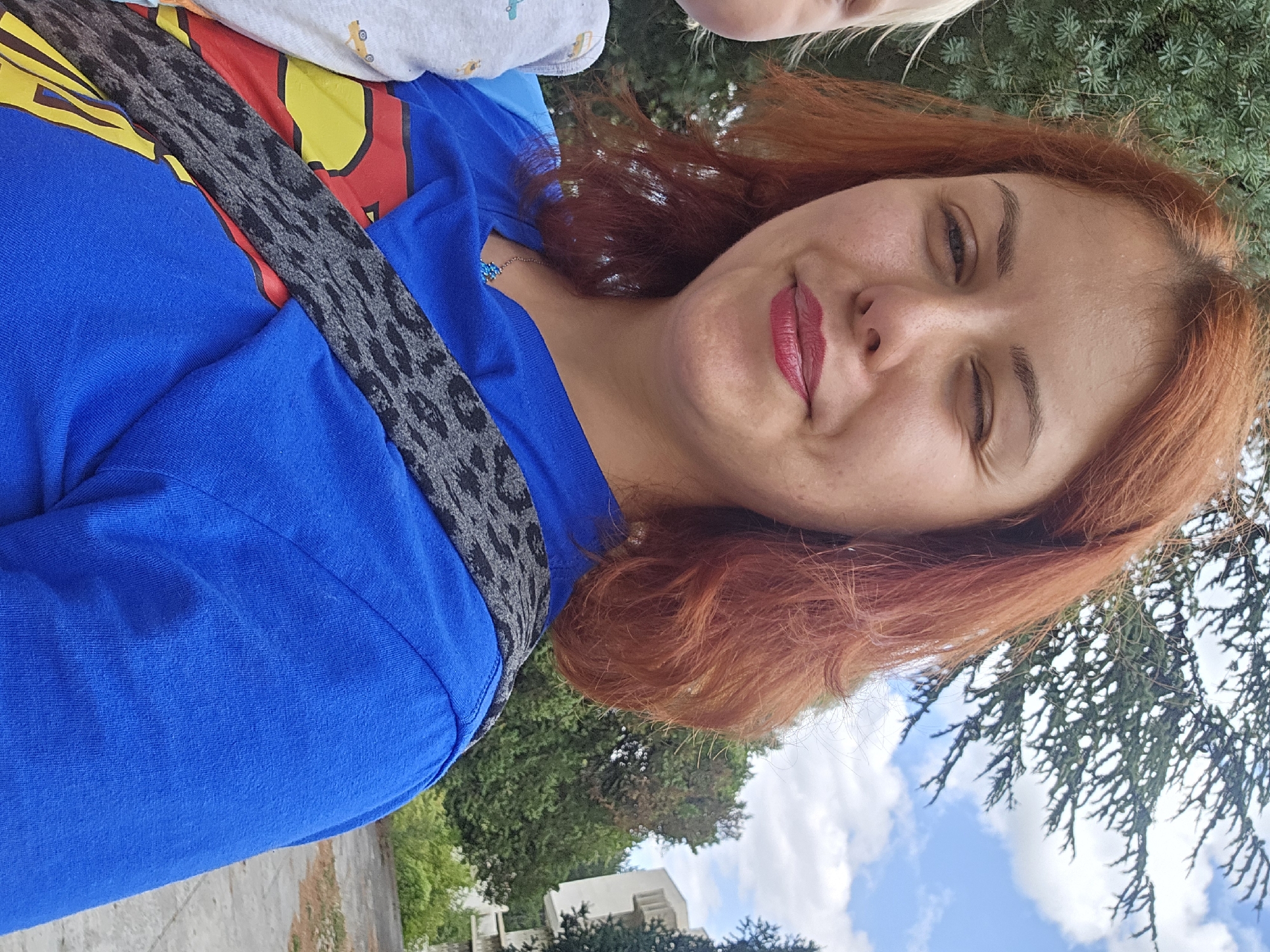 A woman with auburn hair is captured in a close-up, selfie-style shot. She is winking and smiling, with bright red lipstick on her lips. She is wearing a blue t-shirt, which has a red and yellow Superman logo visible underneath a grey leopard-print strap across her shoulder. To her left, a small portion of a child wearing a grey shirt with small yellow car prints is visible. The background consists of green trees and a blue sky with scattered clouds, suggesting an outdoor setting. The woman is wearing a delicate blue necklace with a star-shaped pendant. The overall scene appears casual and happy, possibly taken in a park or residential area.