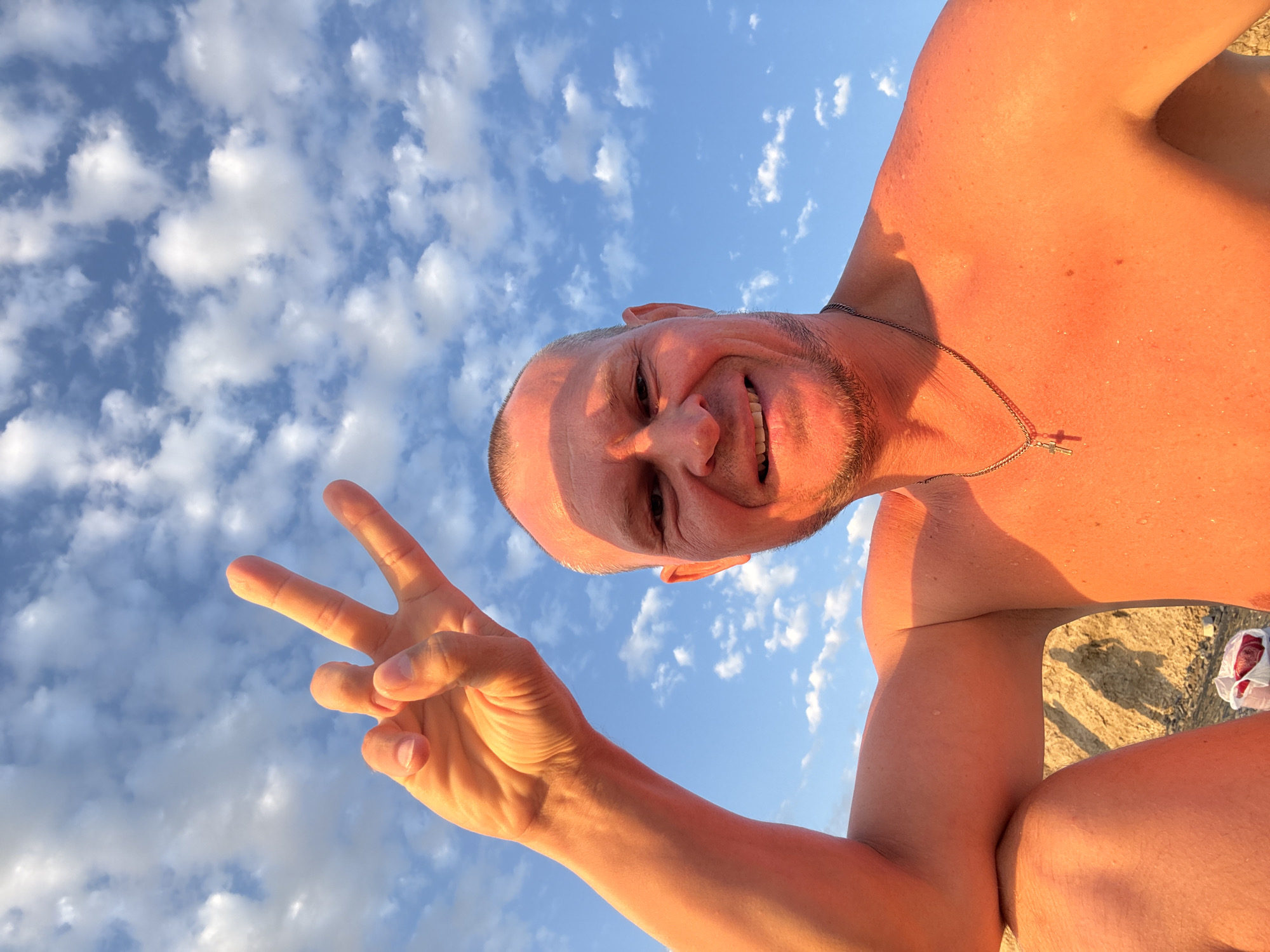 An outdoor, close-up, eye-level, vertical selfie is captured from a low angle, featuring a bald, fair-skinned man in his late 30s or early 40s. He is bare-chested and smiling broadly, with his right arm raised, displaying two fingers in a peace sign against a bright blue sky filled with scattered white clouds. The man has a light beard and is wearing a silver chain with a cross pendant. The setting appears to be a sandy or rocky area, possibly a beach or hillside, with the ground visible in the lower portion of the image. The lighting suggests it is either sunrise or sunset, casting a warm, orange glow on the man's skin. There is no identifiable information about the city of Zharbulak, Kazakhstan in the image.