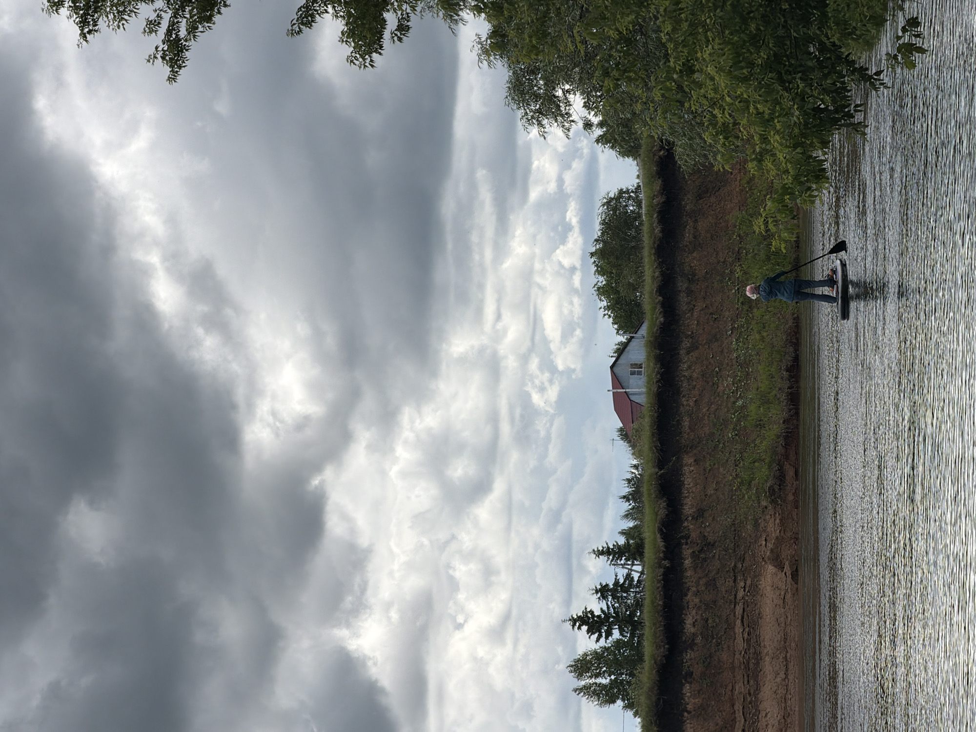 The image depicts a solitary individual paddleboarding on a placid river in Il'inka, Russia, under an overcast sky. The sky, dominating the upper two-thirds of the frame, is filled with dense, uniform grey and white clouds. The river occupies the lower quarter, its surface subtly rippled, reflecting the muted light.
In the mid-ground, a person stands on a dark-colored paddleboard, facing away from the viewer and towards the distant riverbank. The individual is dressed in a dark teal or green jacket, dark pants, and a light-colored head covering, actively propelling the board with a black paddle held on the right side.
Behind the paddleboarder, a steep, eroded riverbank rises from the water. Its lower section reveals layers of reddish-brown earth and sand, while the upper portion is covered with green grass and varied vegetation. Along the top of the bank, a line of trees extends across the scene. On the left, several coniferous trees are visible, while on the right, deciduous trees and bushes are present. Partially obscured by trees on the upper right, a single-story house with a reddish-brown gabled roof and light-colored siding is situated on the elevated land. A portion of a tree with green foliage extends into the foreground on the right, partially framing the scene.