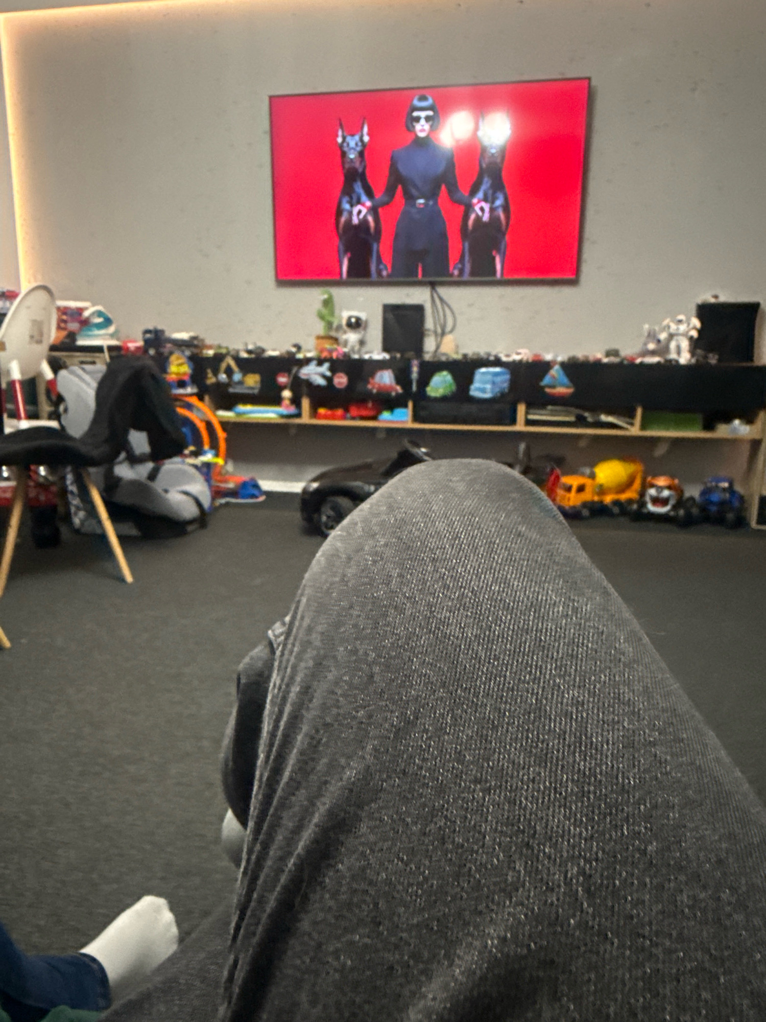 In Gorzów, Poland, a television displays an image of three figures, two dogs and a person in dark attire, against a red background. Below the television, a shelf is laden with various toys, including a toy truck and a miniature cement mixer. In the foreground, a person wearing dark denim jeans sits on a dark carpeted floor, with their legs partially obscuring the view. To the left, a child's white sock and a portion of blue clothing are visible. The room is illuminated by warm lighting originating from the upper left corner, casting a soft glow on the wall and the television screen.