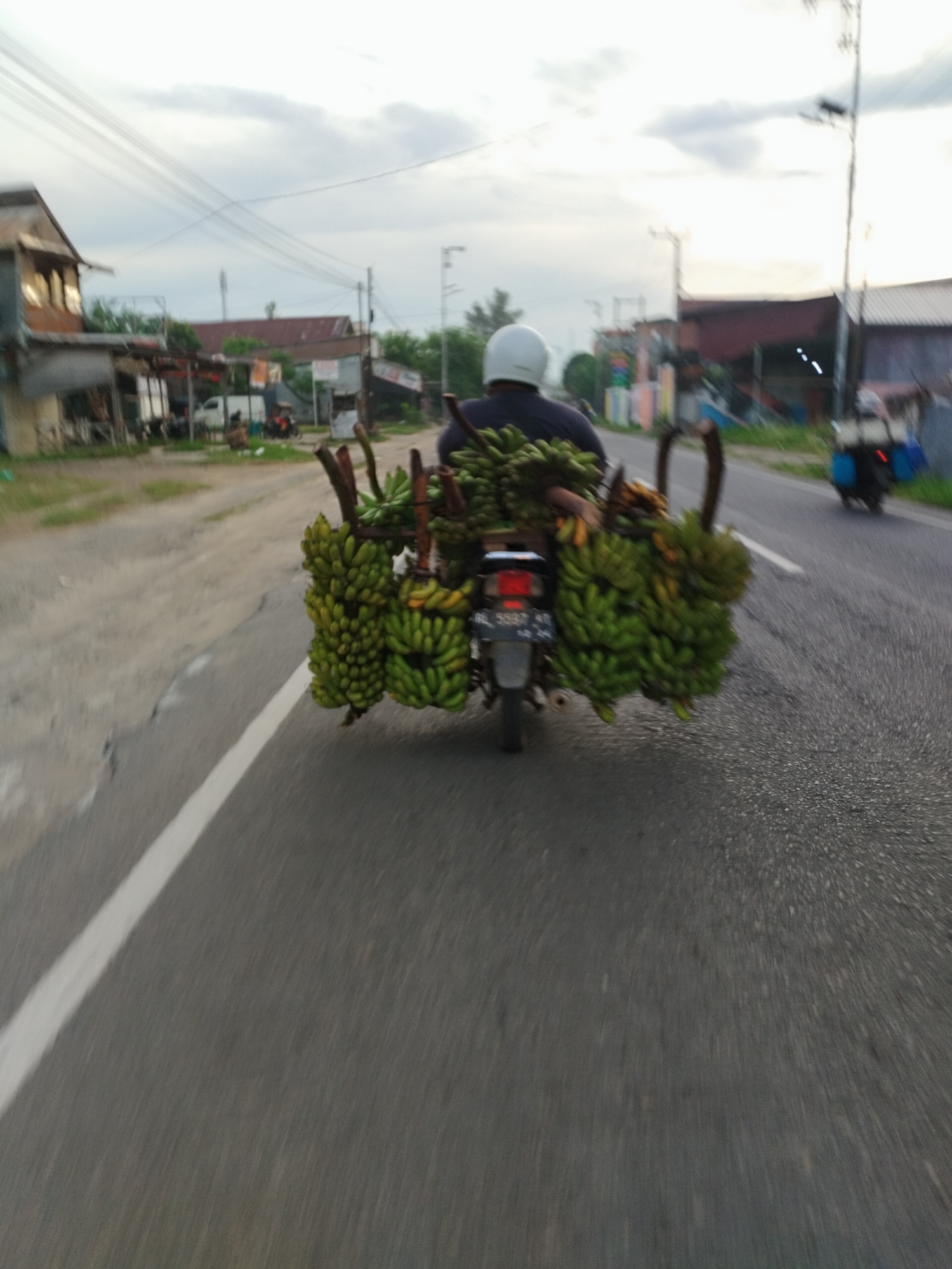 A person wearing a white helmet rides a motorcycle loaded with a large quantity of bananas, predominantly green, on the right side of a paved road. The bananas are arranged in bunches, some extending outwards from the motorcycle. The motorcycle has a license plate visible. The road has a white line marking the edge. In the background, there are buildings with tin roofs and overhead power lines, suggesting a semi-urban or rural setting. The sky is overcast. Another motorcycle is visible in the distance on the right. The image appears to be taken from another moving vehicle.