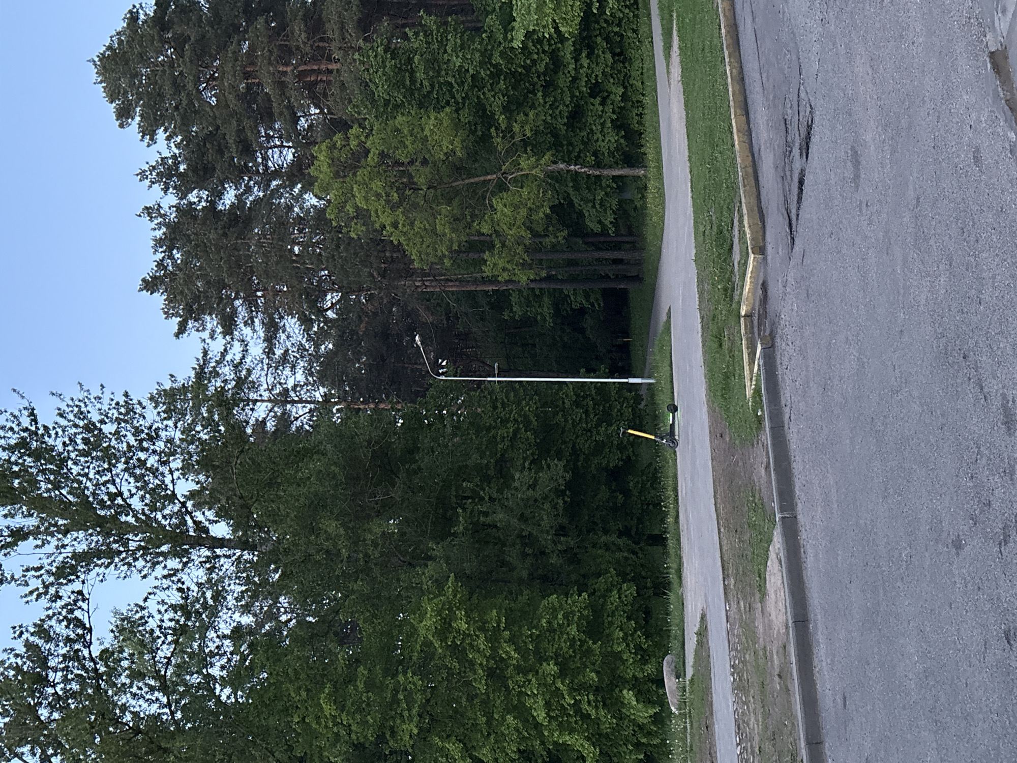 The image presents a sideways view of an urban landscape, likely a street or park area in Minsk, Belarus, based on the environment typical for that region. A paved road occupies the right side of the frame, showing signs of wear and tear. Adjacent to the road is a pathway, bordered by grass and trees. A yellow and black scooter lies near the path. A street light is centrally located. The sky is visible at the top left corner. The overall atmosphere suggests a quiet, possibly residential area within the city.