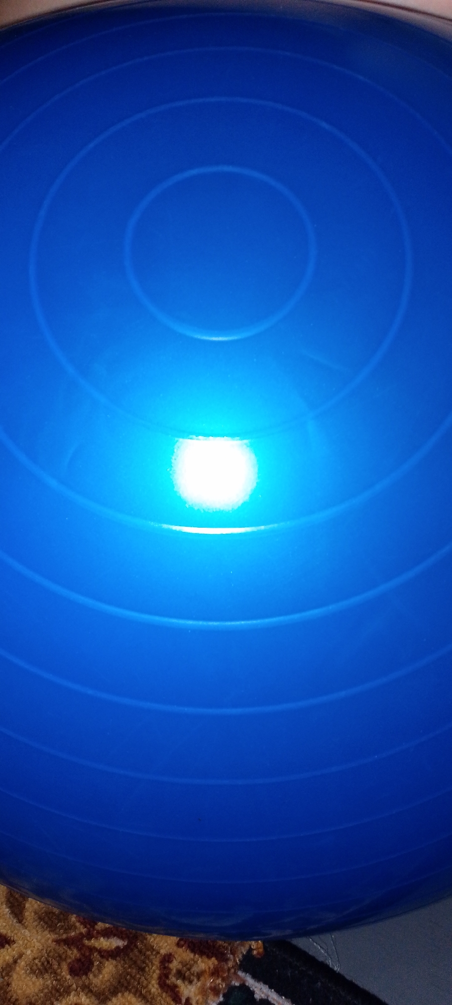 A bright, circular light source reflects off a smooth, blue exercise ball. The ball has several concentric circles embossed on its surface, creating a textured appearance. The bottom portion of the ball rests on a patterned rug. The scene is lit by an unseen light source that creates a strong highlight on the ball.
