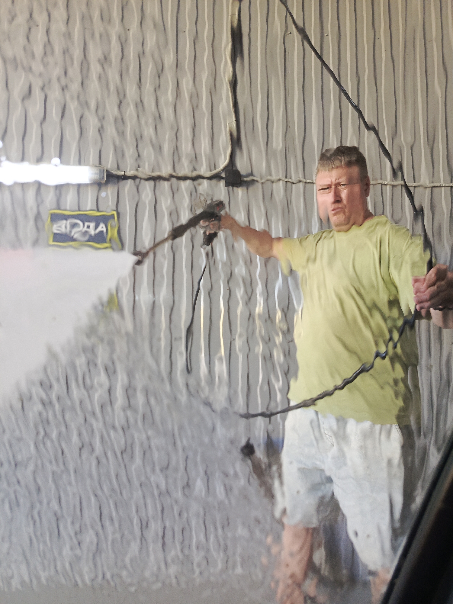 A middle-aged man is washing a car, possibly at a self-service car wash in Chaikovsky, Russia. The man is wearing a lime green T-shirt and light-colored shorts. He is holding a pressure washer wand and spraying the car with water. The word "ВОДА" (water) is visible on a sign in the background. The image appears to be taken from inside the car being washed.