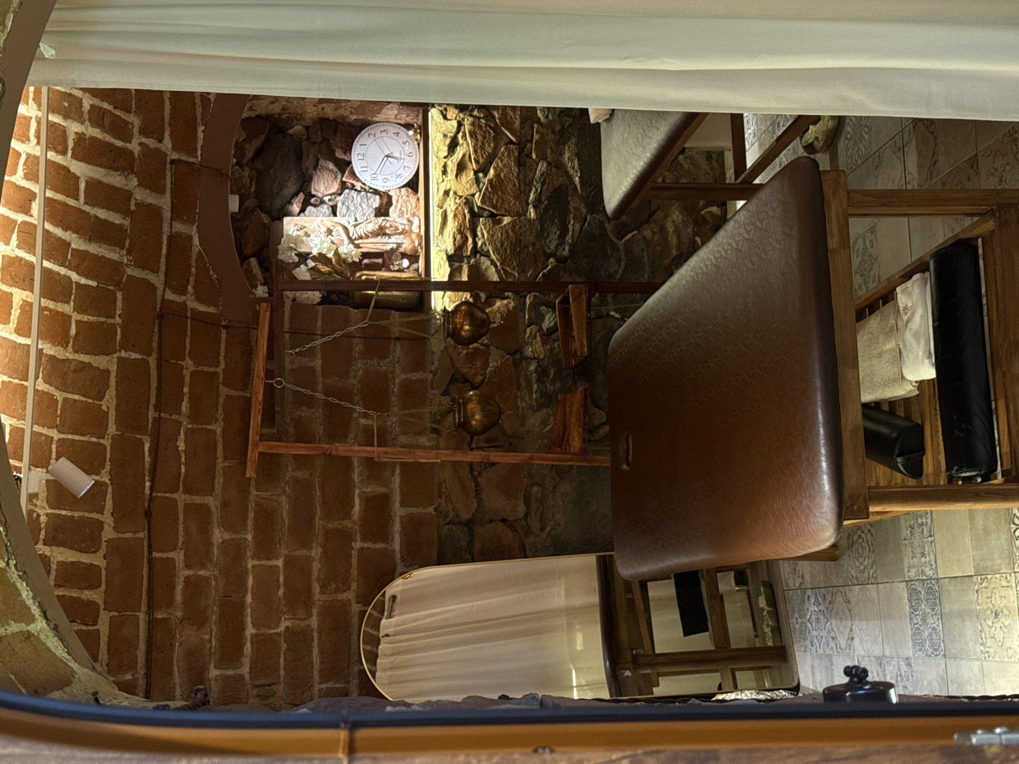This image appears to be an interior shot of a room with distinctive architectural features, potentially located in Yerevan, Armenia, given the building style. The room's walls are constructed of red brick on the left side and a combination of brick and stone on the right. A circular window or opening is visible on the left, partially obscured by the brick wall. A vintage-style wall clock is mounted on the wall made of stone, above a wooden shelf adorned with decorative objects. A wooden rack with what appears to be towels or linens is positioned towards the right side of the image. A light-colored curtain or fabric hangs from the ceiling. The scene suggests a unique architectural style, possibly a renovated space that retains historical elements.