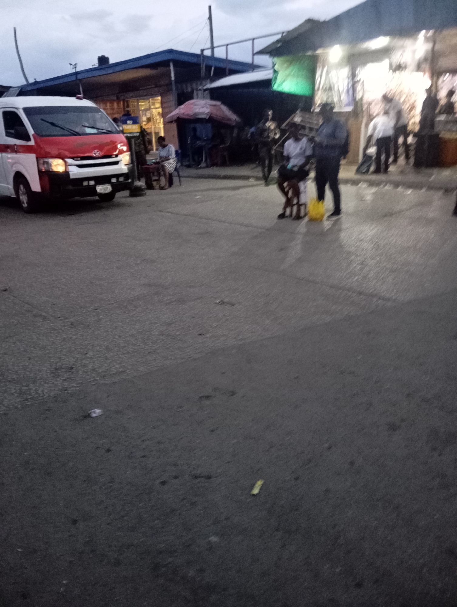 The image depicts a street scene in Port Harcourt, Nigeria, likely during twilight or overcast conditions. A white and red van, possibly an ambulance or commercial vehicle, is parked on the left side of the asphalt road, facing left. Its headlights are on. A "NO PARKING" sign is visible near the van. In the background, a building with a blue roof and what appears to be shops or stalls is visible. Several people are present: a person is sitting on a chair near the building, and other individuals are standing or walking in front of the shops. One person is sitting on a chair with a yellow bag on the ground beside them, while another person stands nearby holding a yellow object. The overall environment suggests a busy commercial area.